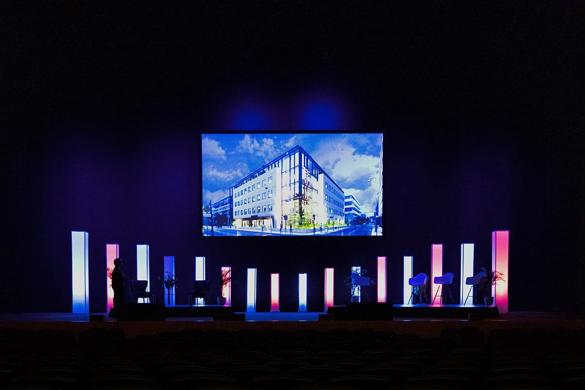 An empty stage with a large screen showing a building, with vertical light columns in pink and blue, and a person setting up chairs.