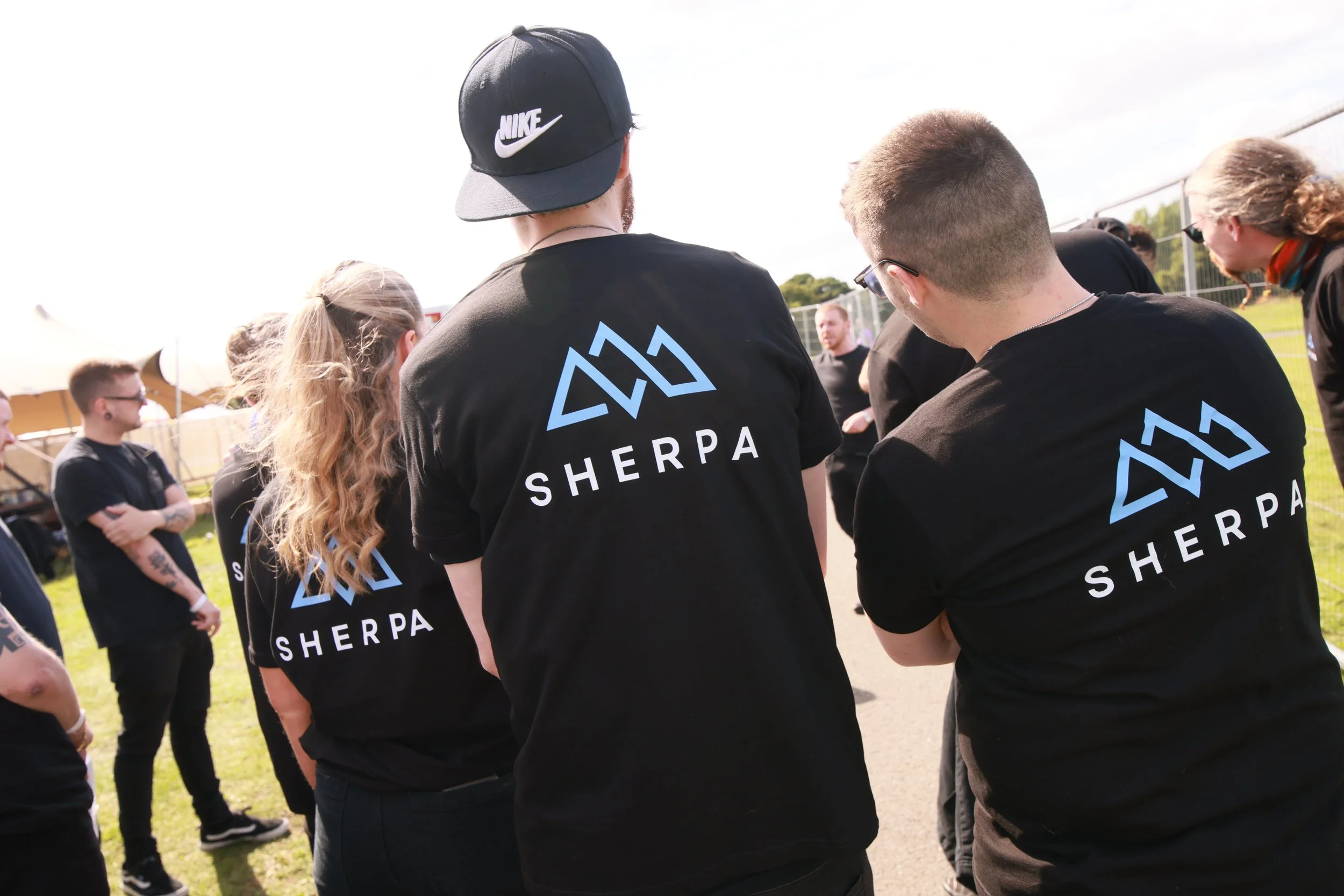 Group of people wearing black T-shirts with 'SHERPA' logo, gathered outdoors on a sunny day, some engaging in conversation, with a fence and green trees in the background.