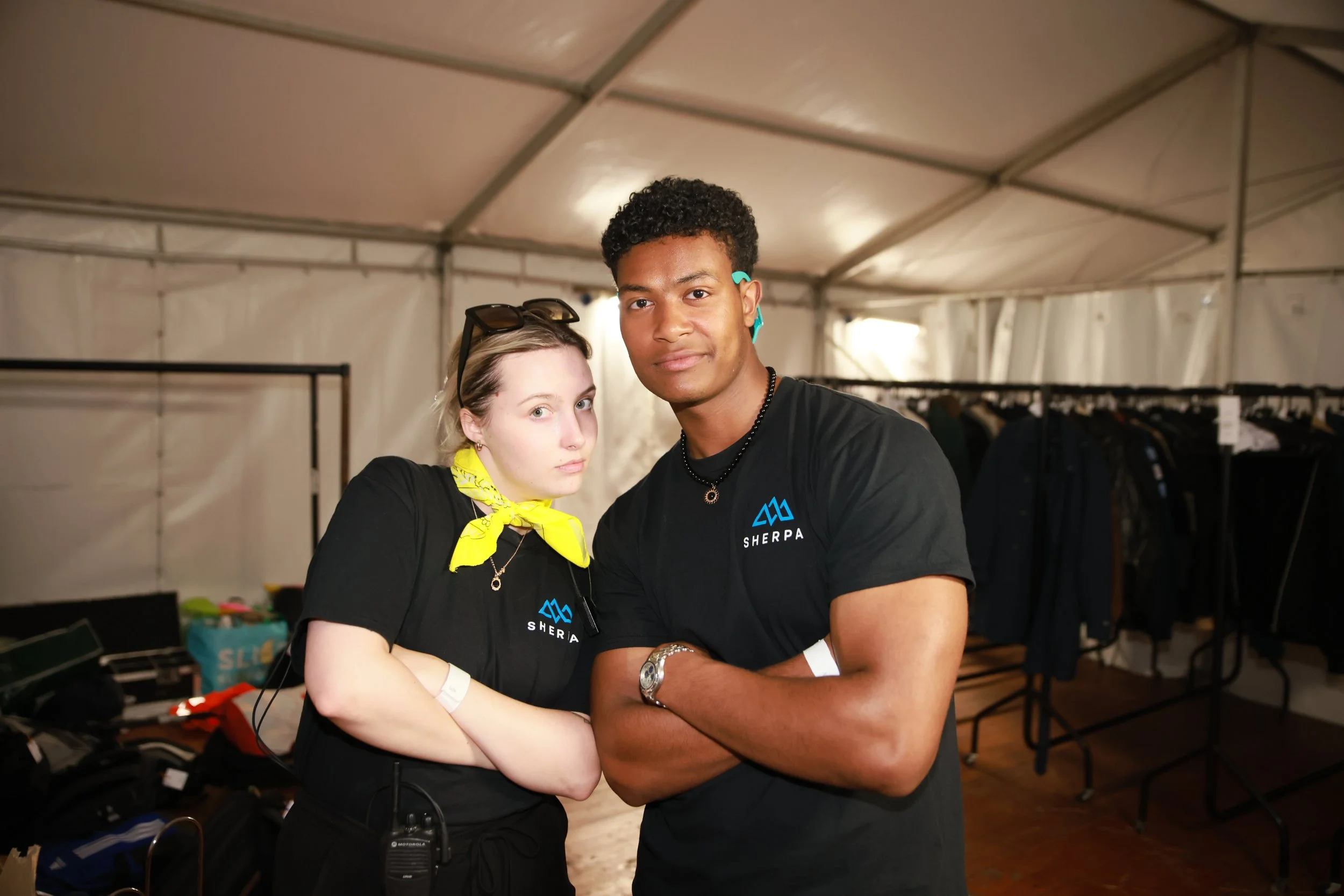 A young woman with blonde hair and a yellow neckerchief draped around her neck, standing with a young man with dark curly hair, both wearing black shirts with a blue mountain logo and the word 'SHERPA.' They are inside a large tent, with clothing racks and various items in the background.