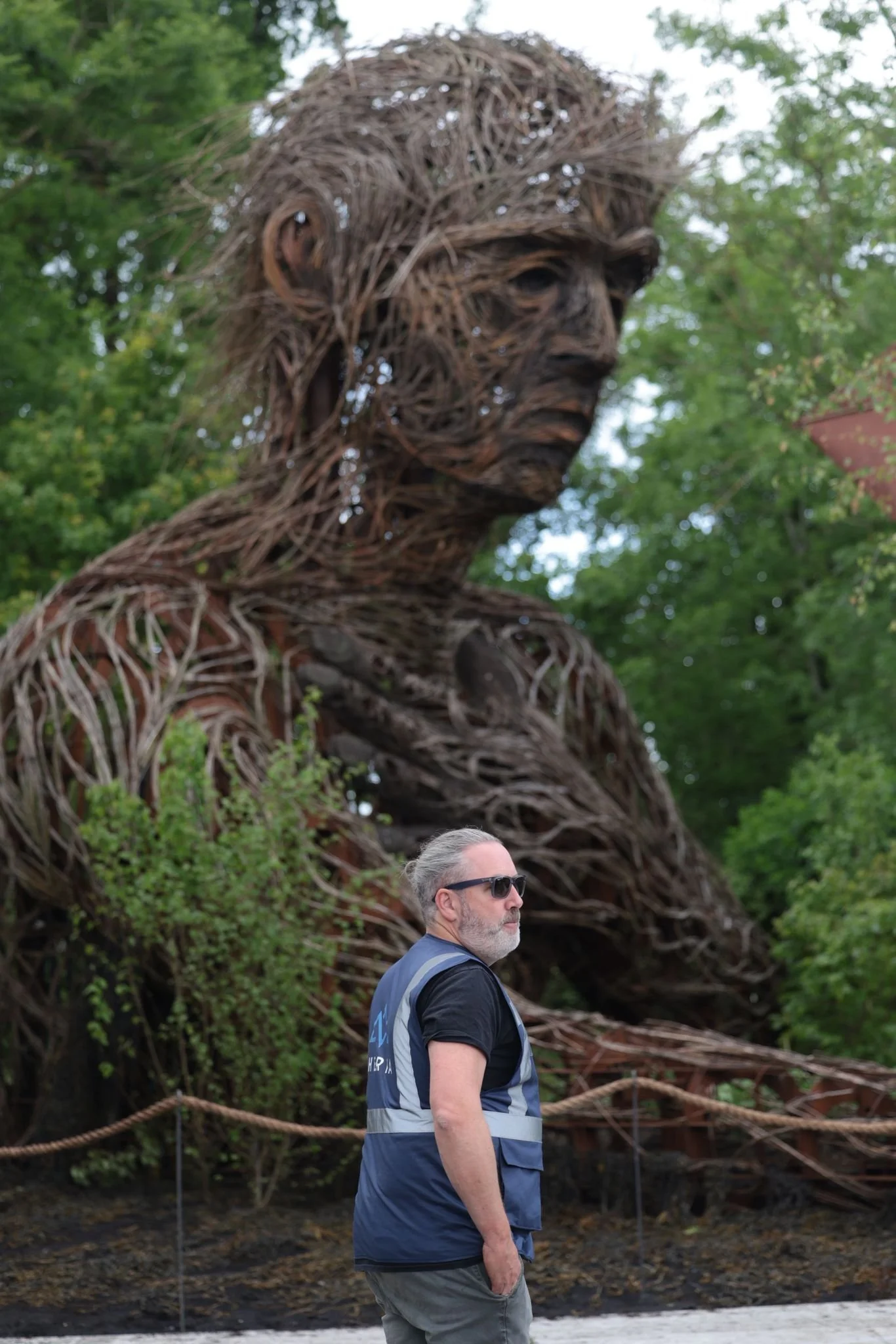 A man with gray hair, beard, wearing sunglasses, a dark T-shirt, and a blue vest stands outdoors. Behind him is a large sculpture of a person's face made from intertwined tree branches and twigs, set in a wooded area with green leaves.