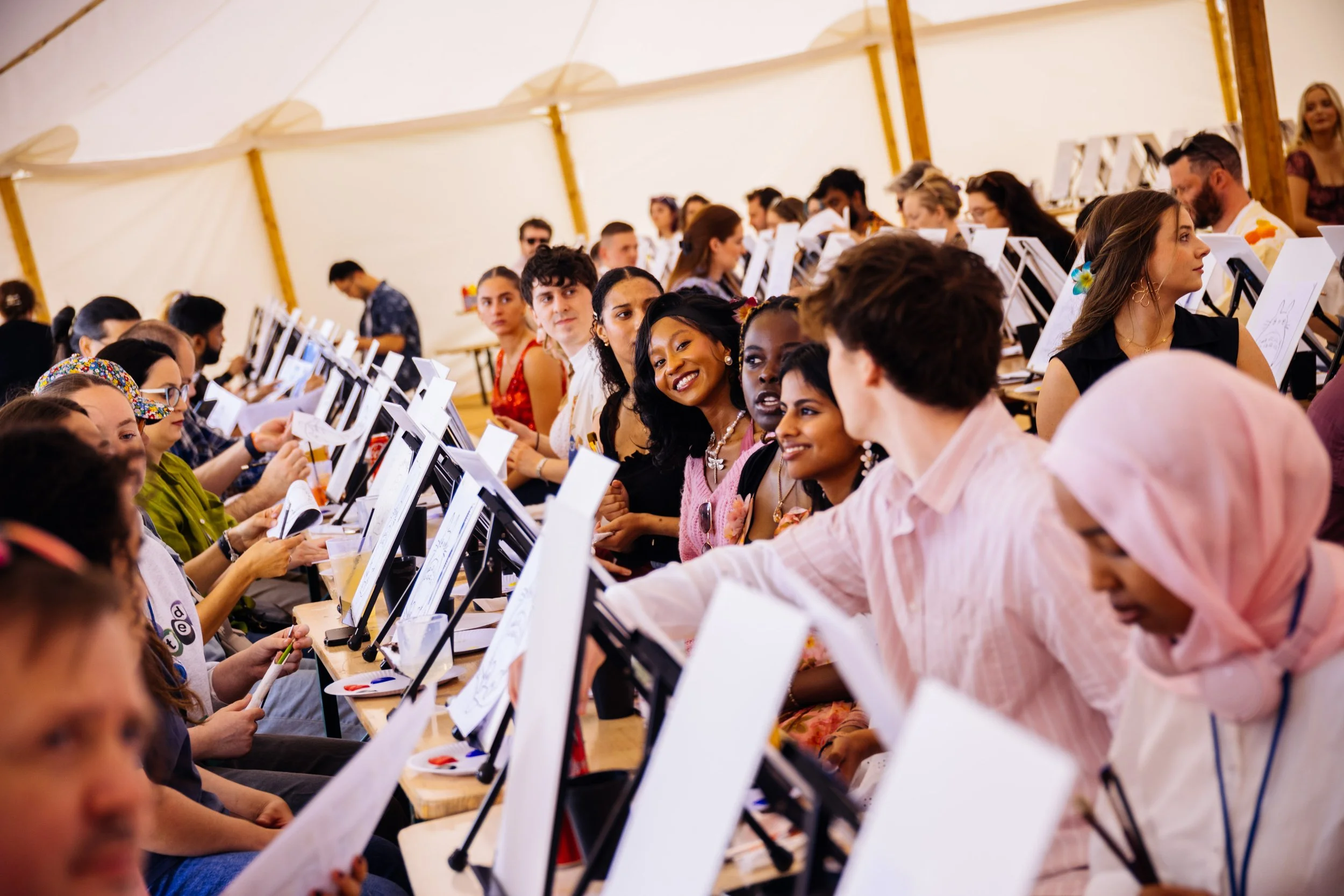 Group of diverse adults sitting at tables in a large tent, engaging in a painting activity with canvases and paints.