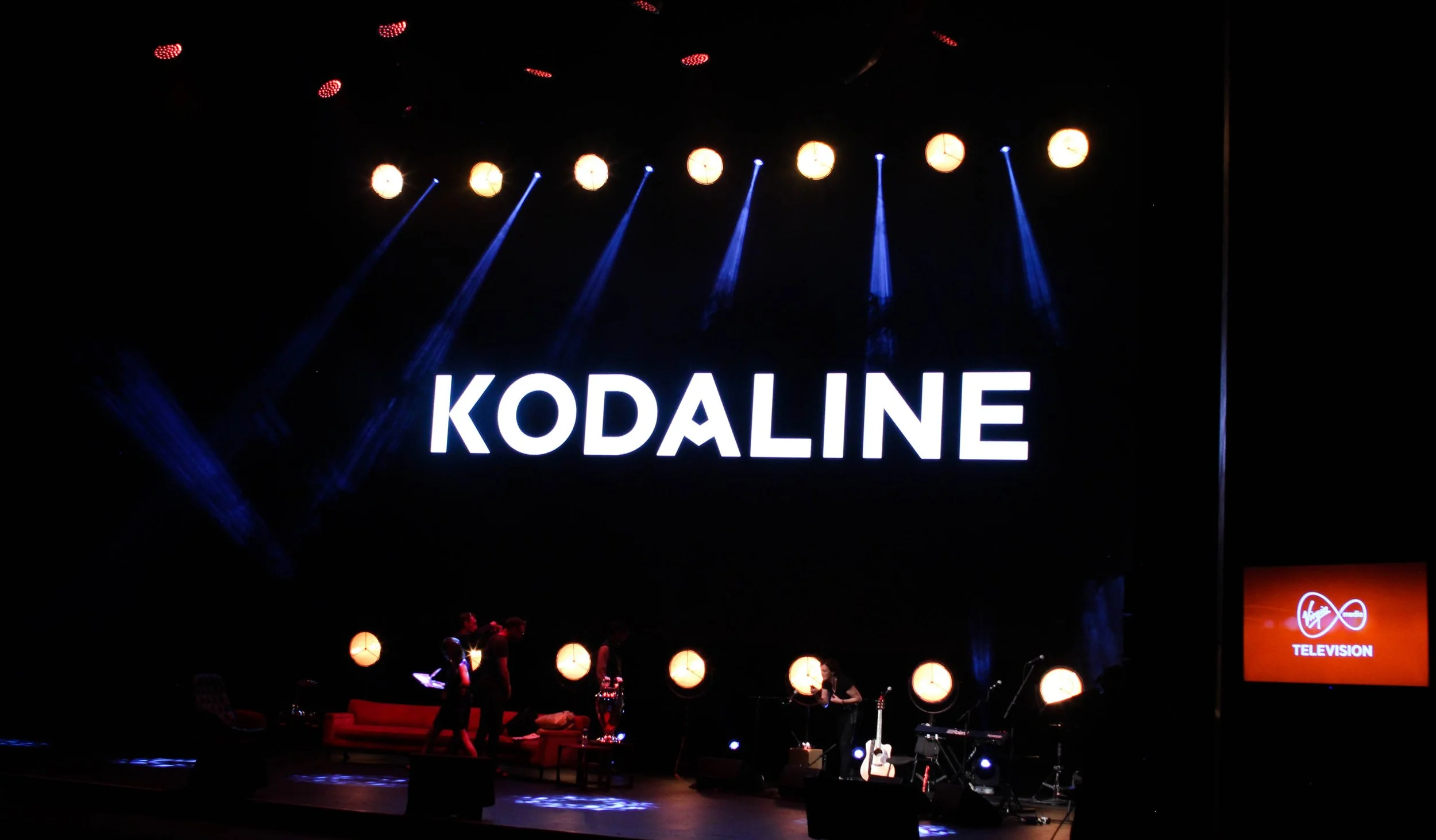 Stage setup with the word 'KODALINE' illuminated in large white letters on a black background, with yellow stage lights hanging overhead and people preparing for a performance.