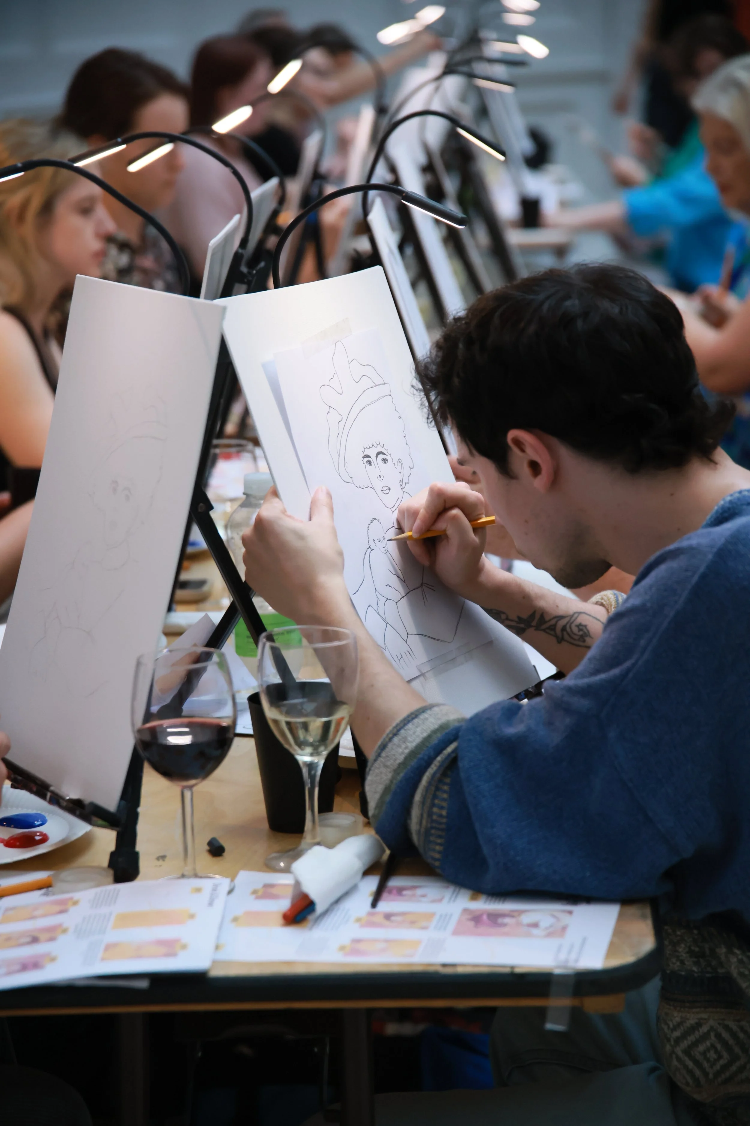 People sitting at a long table drawing and coloring portraits in an art class, with sketchbooks, drawing supplies, and glasses of wine on the table.