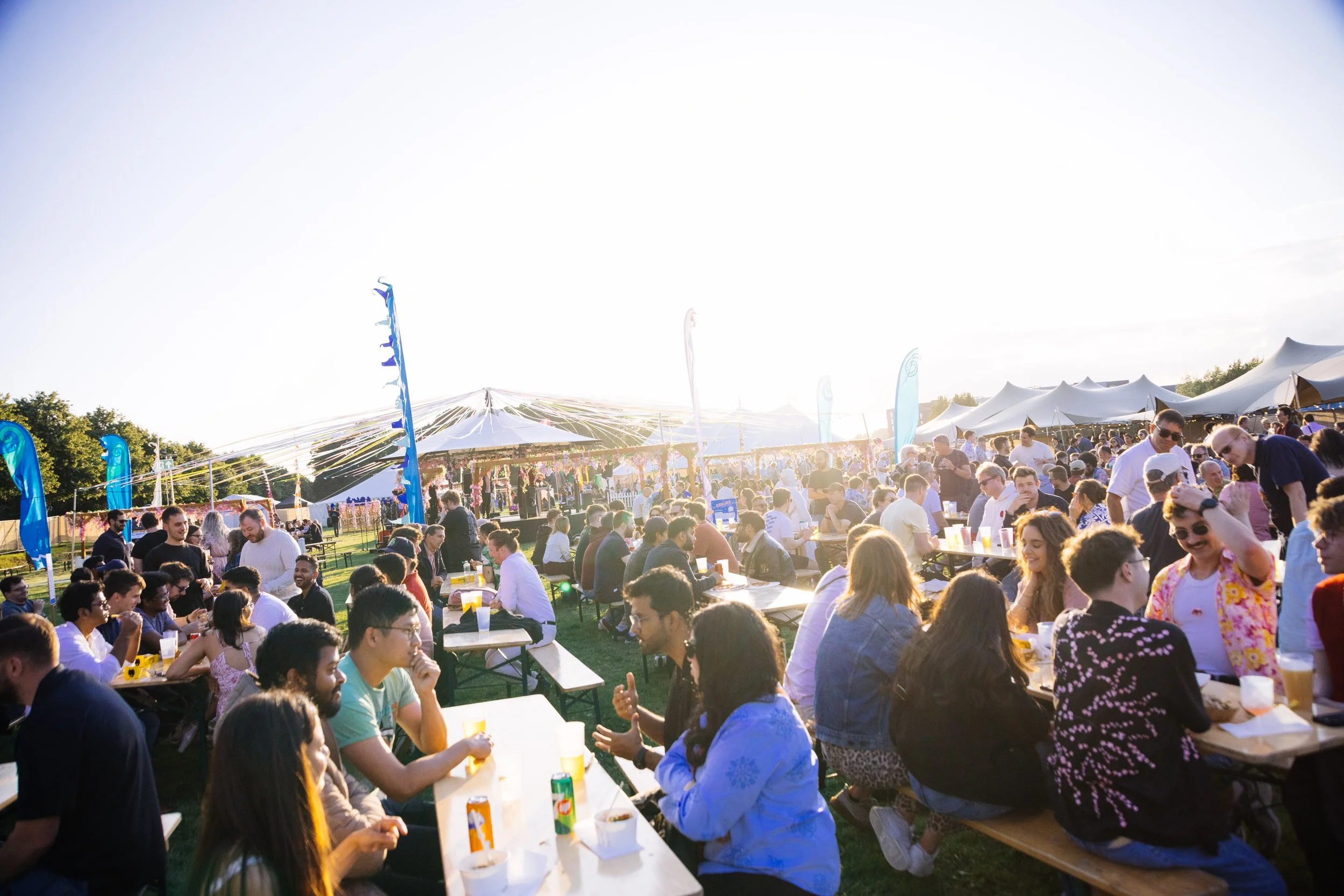 A large crowd of people sitting at long tables outdoors at a festival, with music tents, colorful flags, and clear sky in the background.