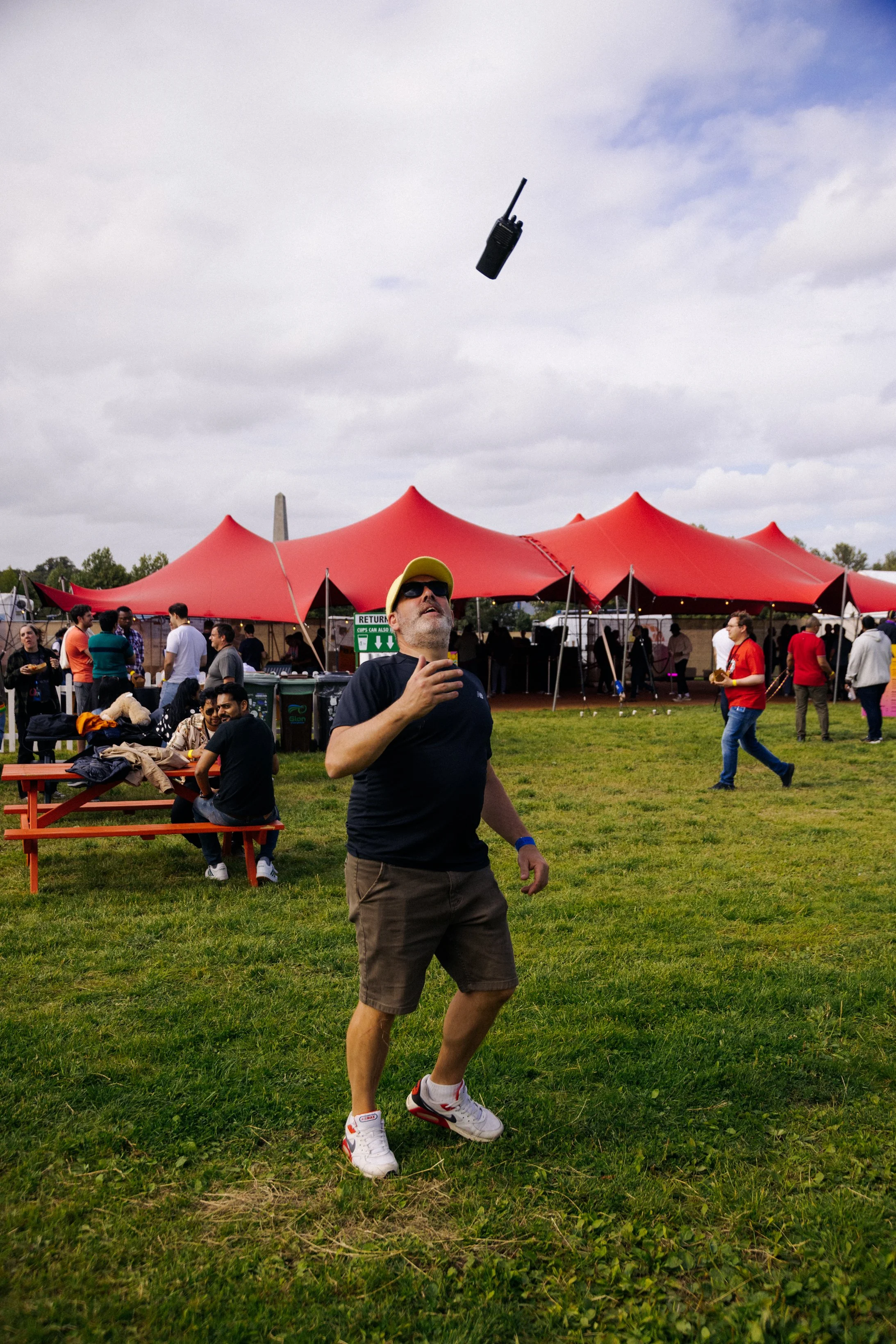 A man wearing sunglasses, a yellow cap, black T-shirt, brown shorts, and white sneakers with red accents is holding a drink and walking on a grassy area at an outdoor event. In the background, there are people, tents with red tops, and a cloudy sky.