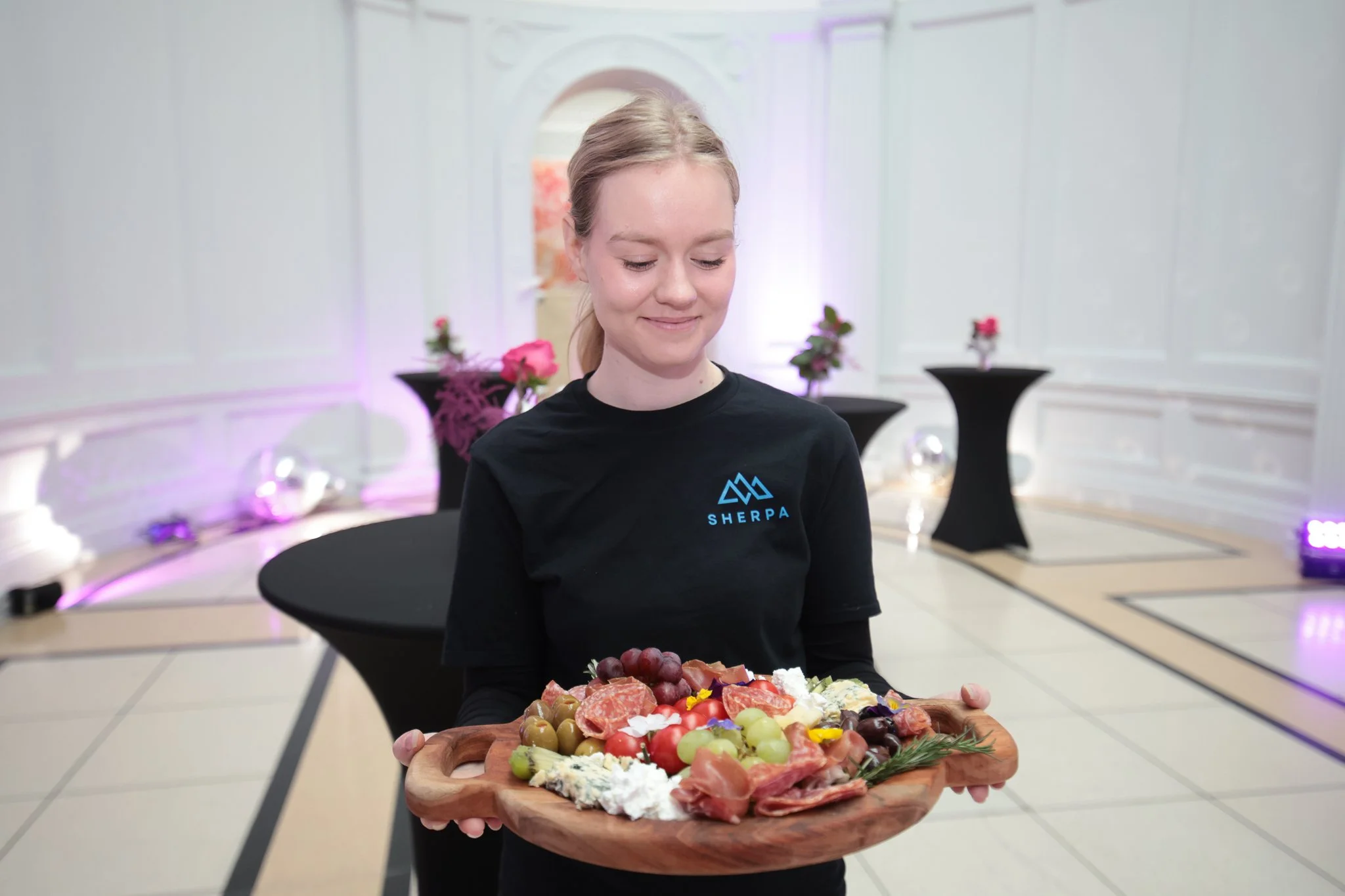 A young woman in a black shirt with 'Sherpa' logo holds a wooden tray of assorted fruits and cheeses at a decorated event space with white walls and floral arrangements in the background.