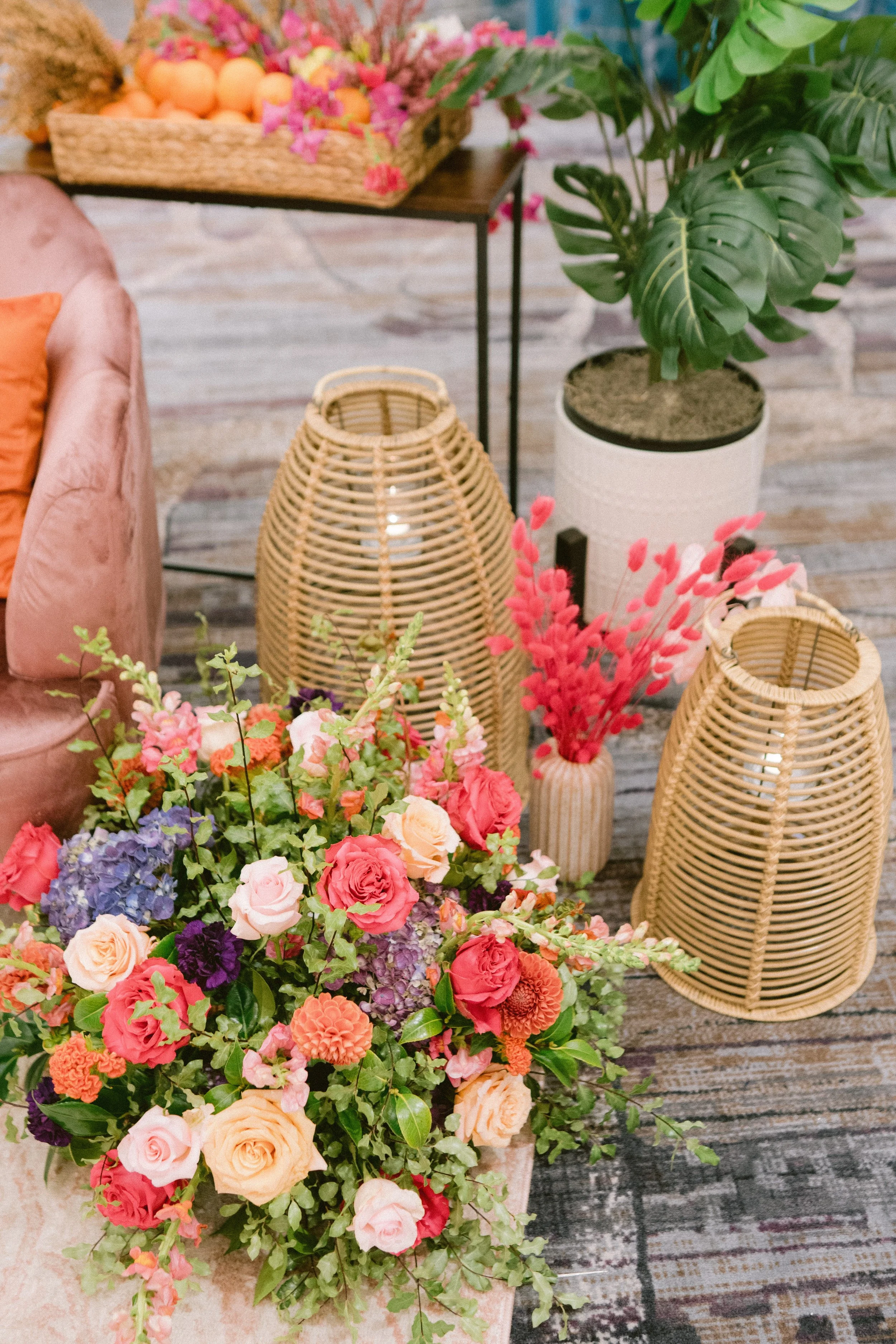 A large bouquet of colorful flowers including roses, hydrangeas, and dahlias in the foreground, with woven rattan lanterns, green potted plant, and a wicker basket filled with eggs and pink flowers on a wooden table in the background.