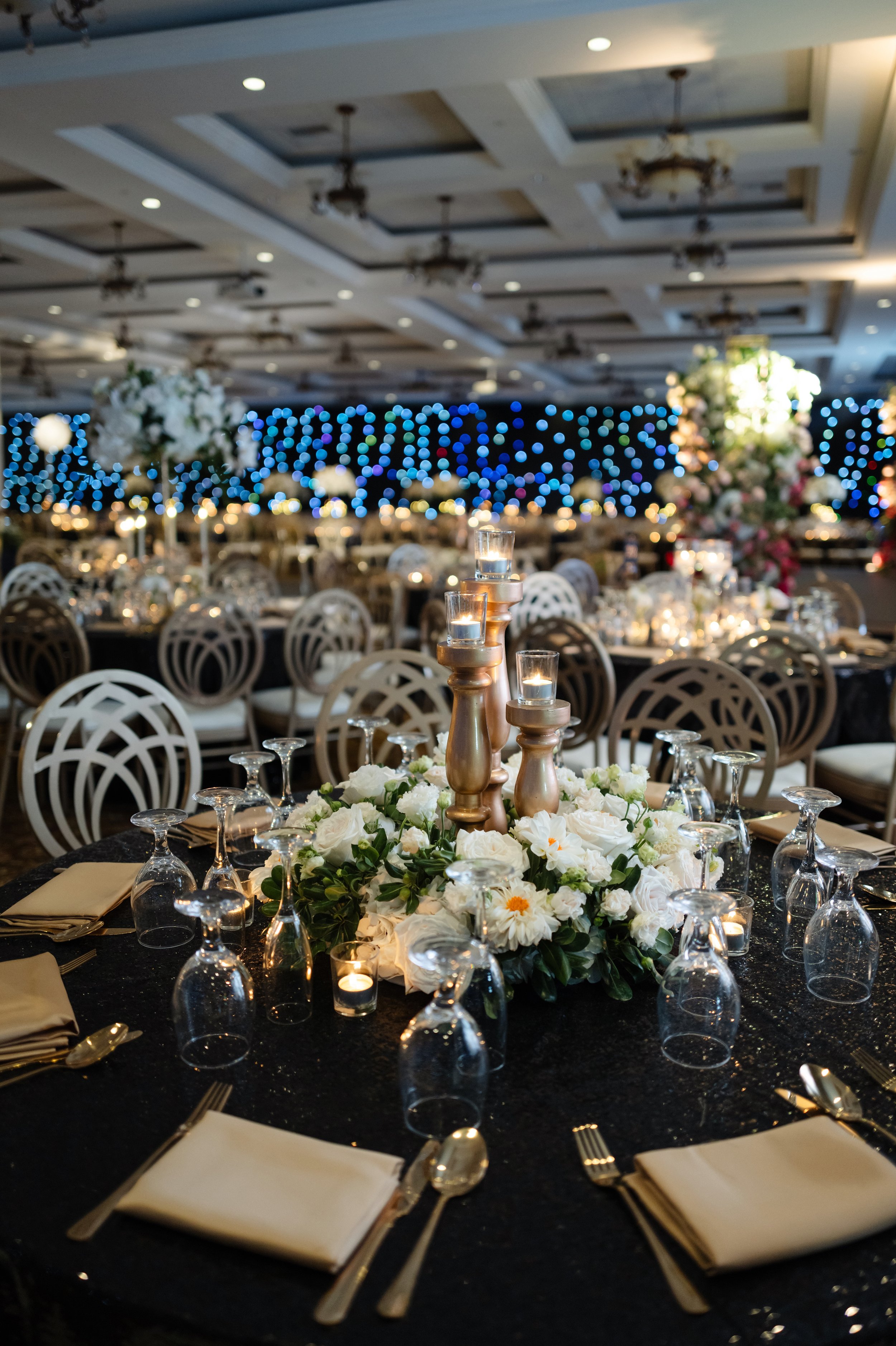 Elegant banquet hall decorated for a special event with round tables, floral centerpieces, candles, and string lights with the words "Congratulations" in the background.