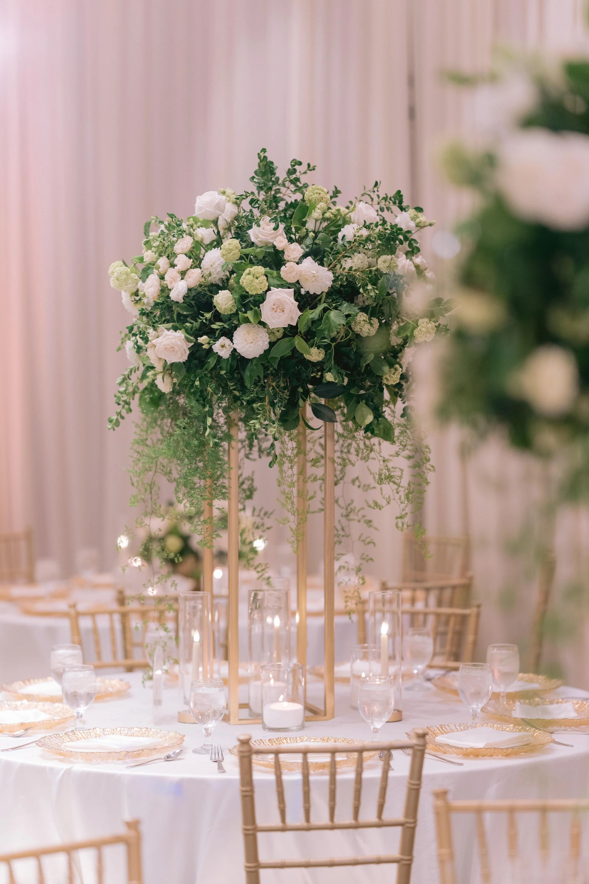 Elegant dining table with a tall centerpiece of white roses, greenery, and candles, set with gold-rimmed plates and glassware at a wedding or event.