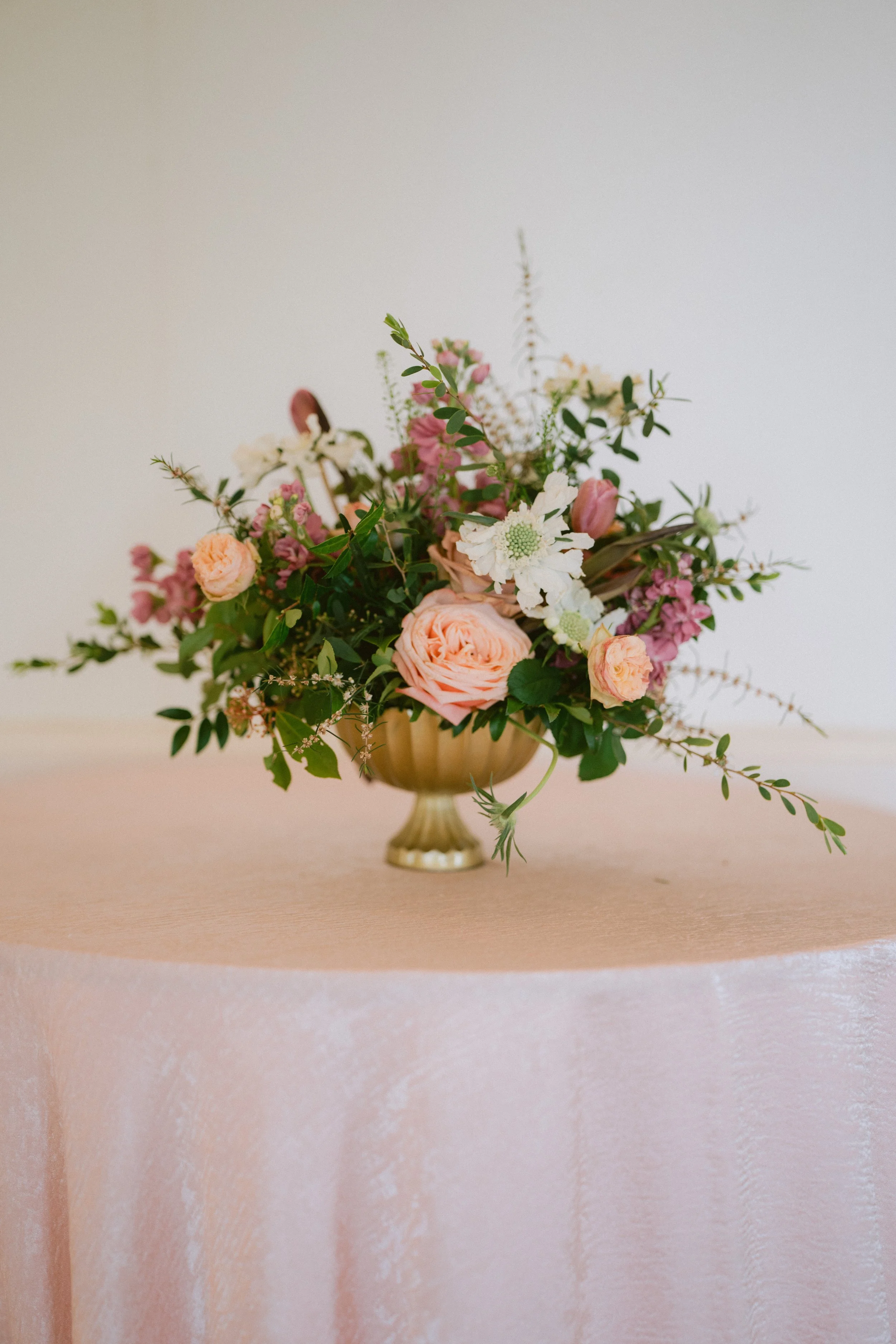 A bouquet of pink, white, and peach flowers arranged in a golden vase on a pink textured table with a pink wall in the background.