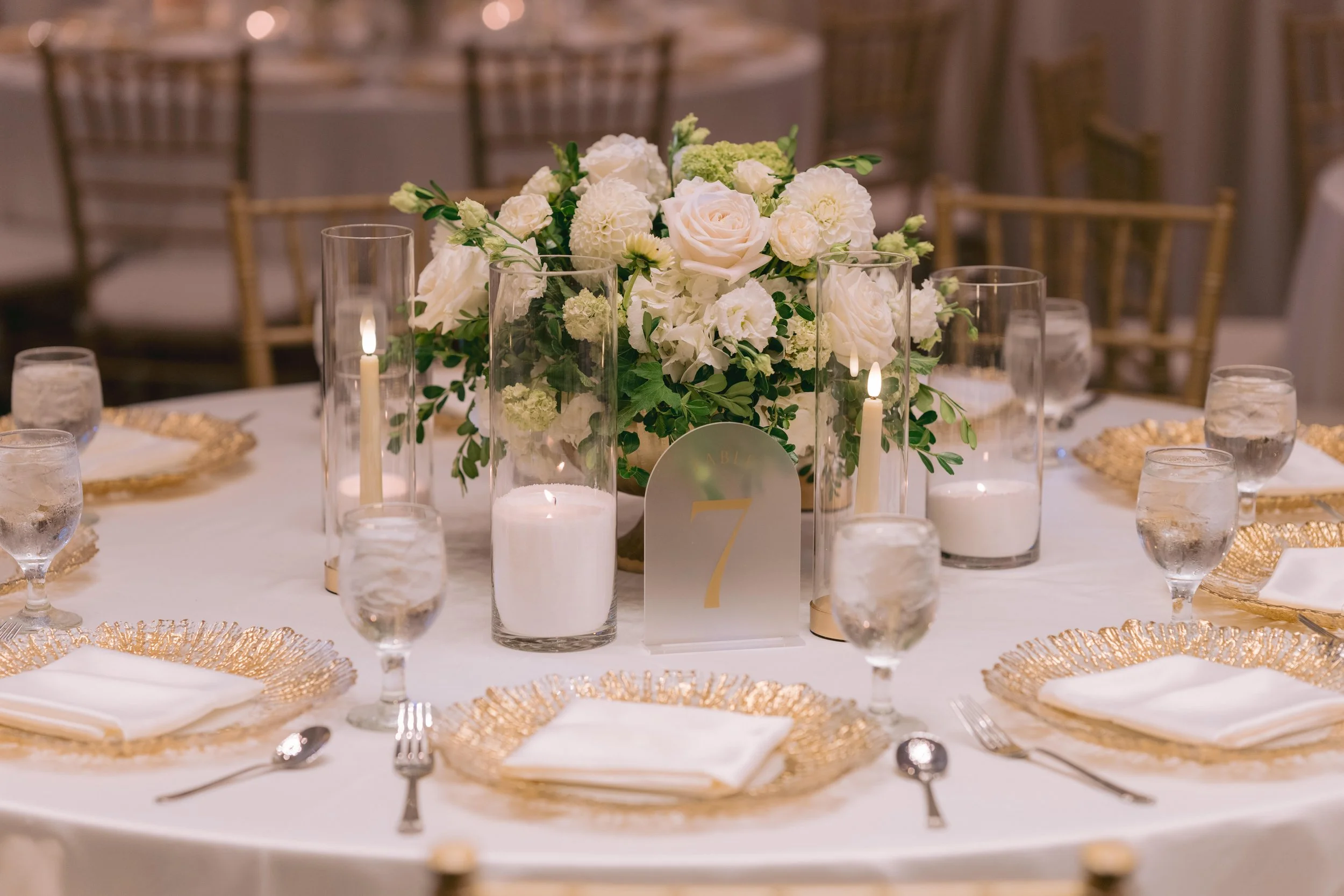 Table centerpiece with white flowers in a vase, surrounded by tall glass candles and smaller votive candles, set on a white tablecloth with gold accents and place settings.