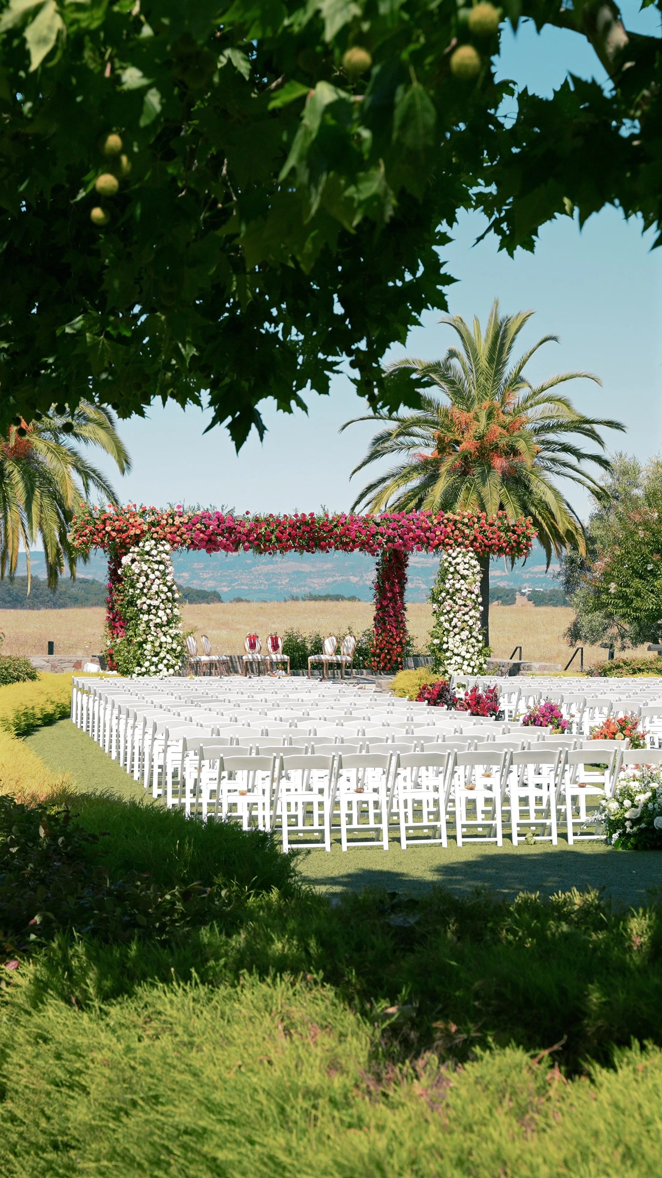 Outdoor wedding ceremony setup with white chairs, a floral arch with pink, red, and white flowers, and tropical trees including palm trees, set in a scenic rural area.