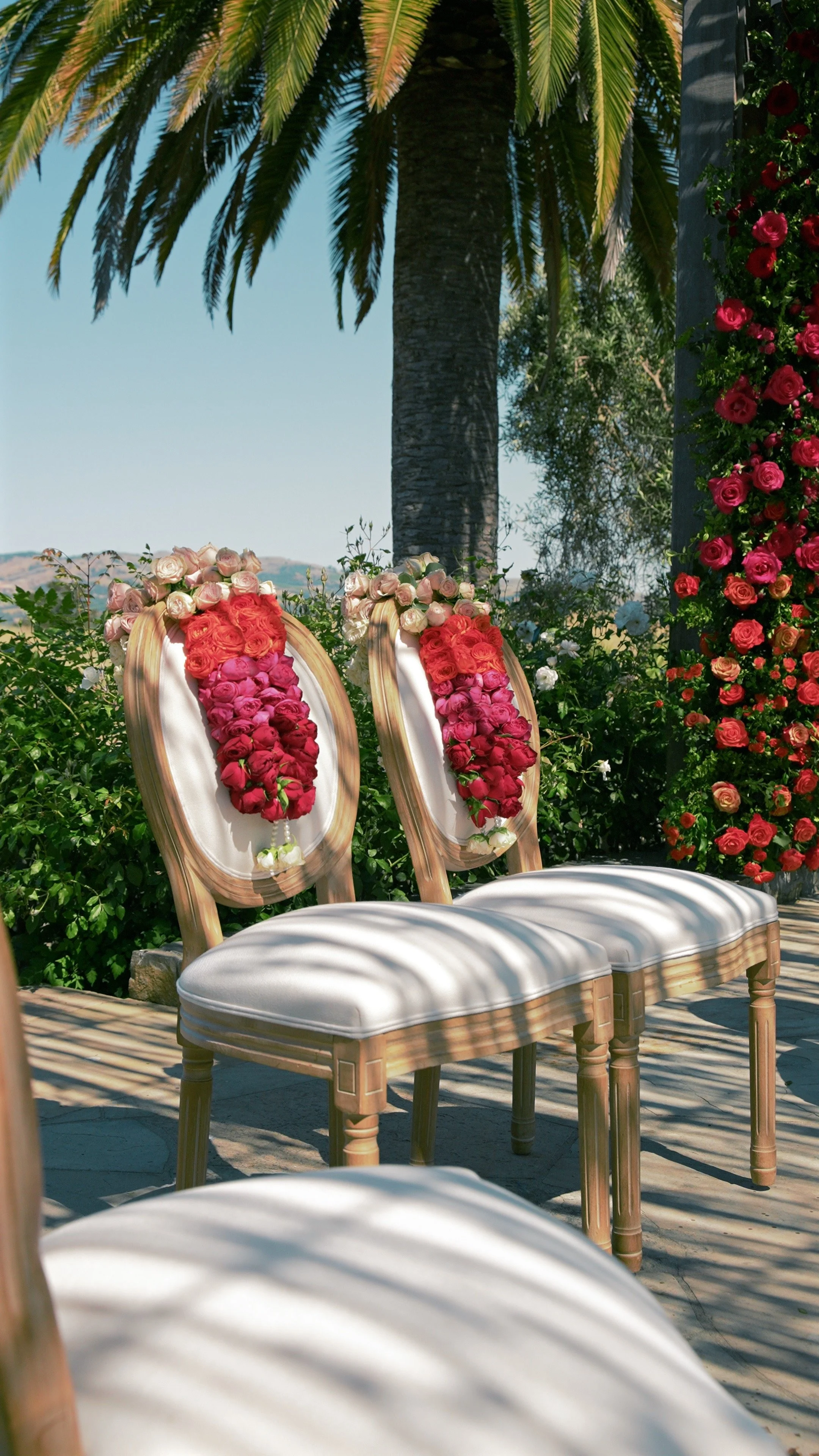 Two chairs decorated with floral designs, set outdoors under a pine tree, with a backdrop of flowering plants and a clear sky, suggesting a wedding or outdoor event.