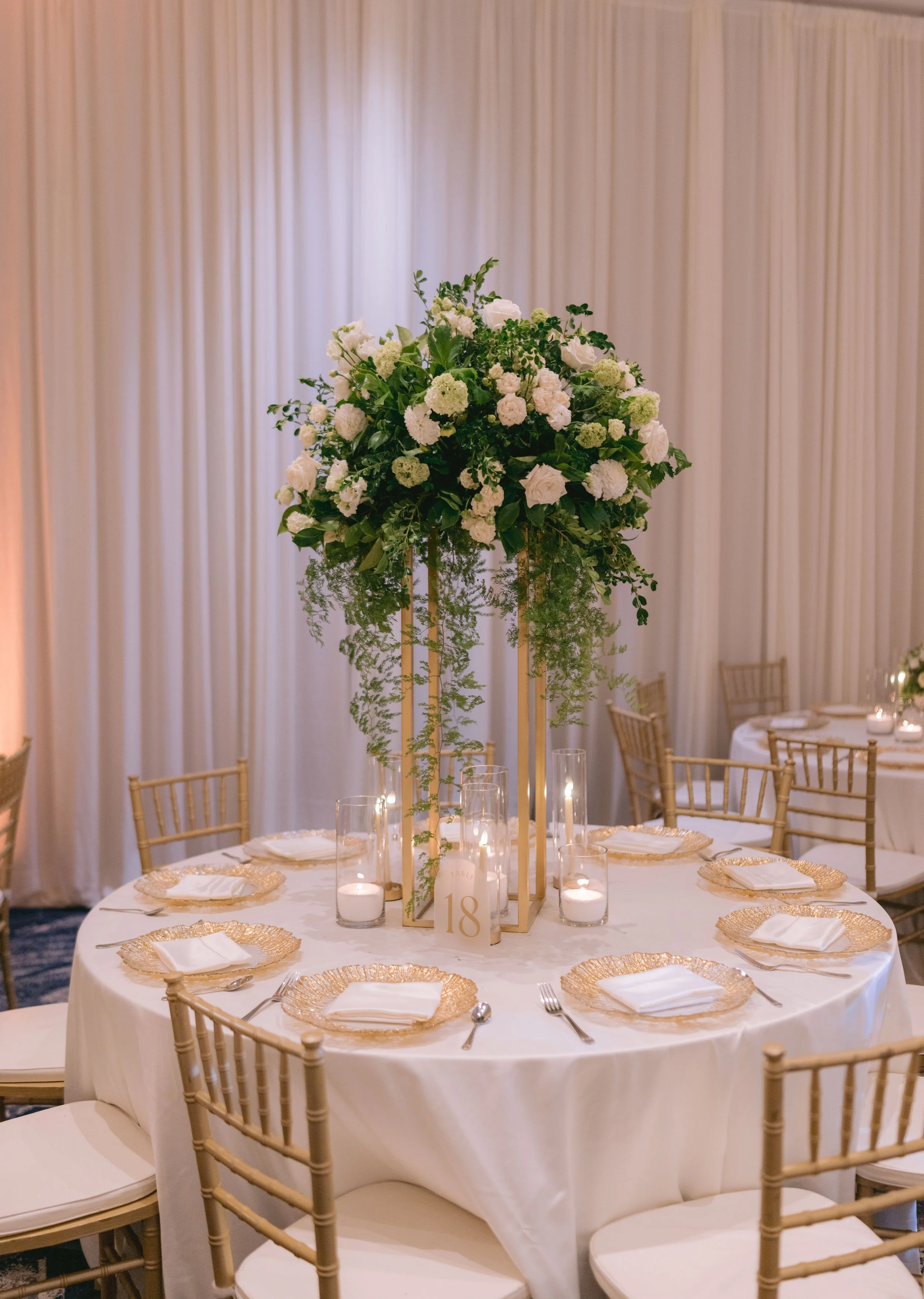 Elegant round wedding reception table with a tall floral centerpiece of white and green flowers, surrounded by candles, gold charger plates, and white napkins, set against a backdrop of cream-colored draped curtains.