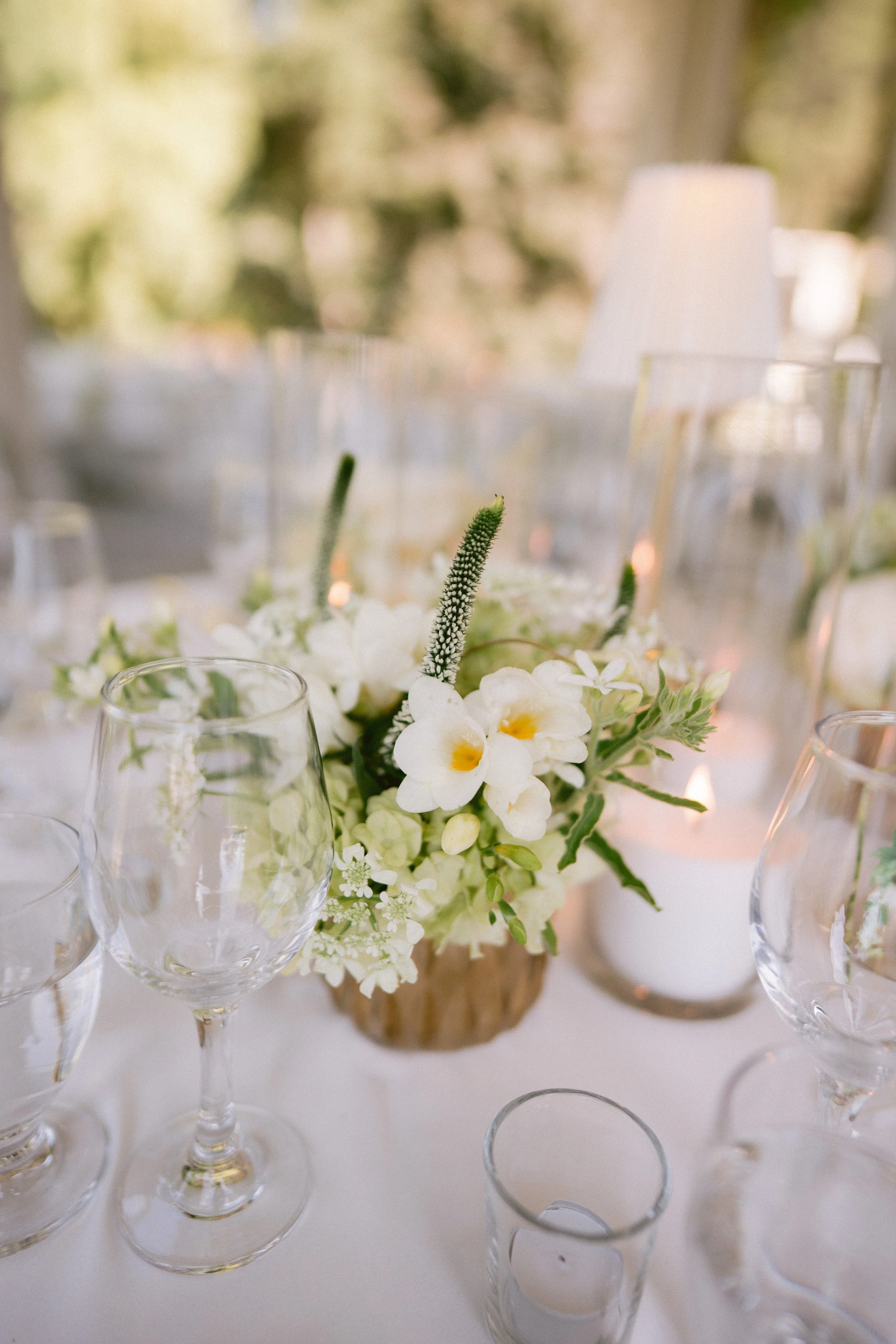 A floral centerpiece with white flowers and green foliage in a brown pot, surrounded by clear glassware and small candles on a white tablecloth, with an out-of-focus outdoor background.