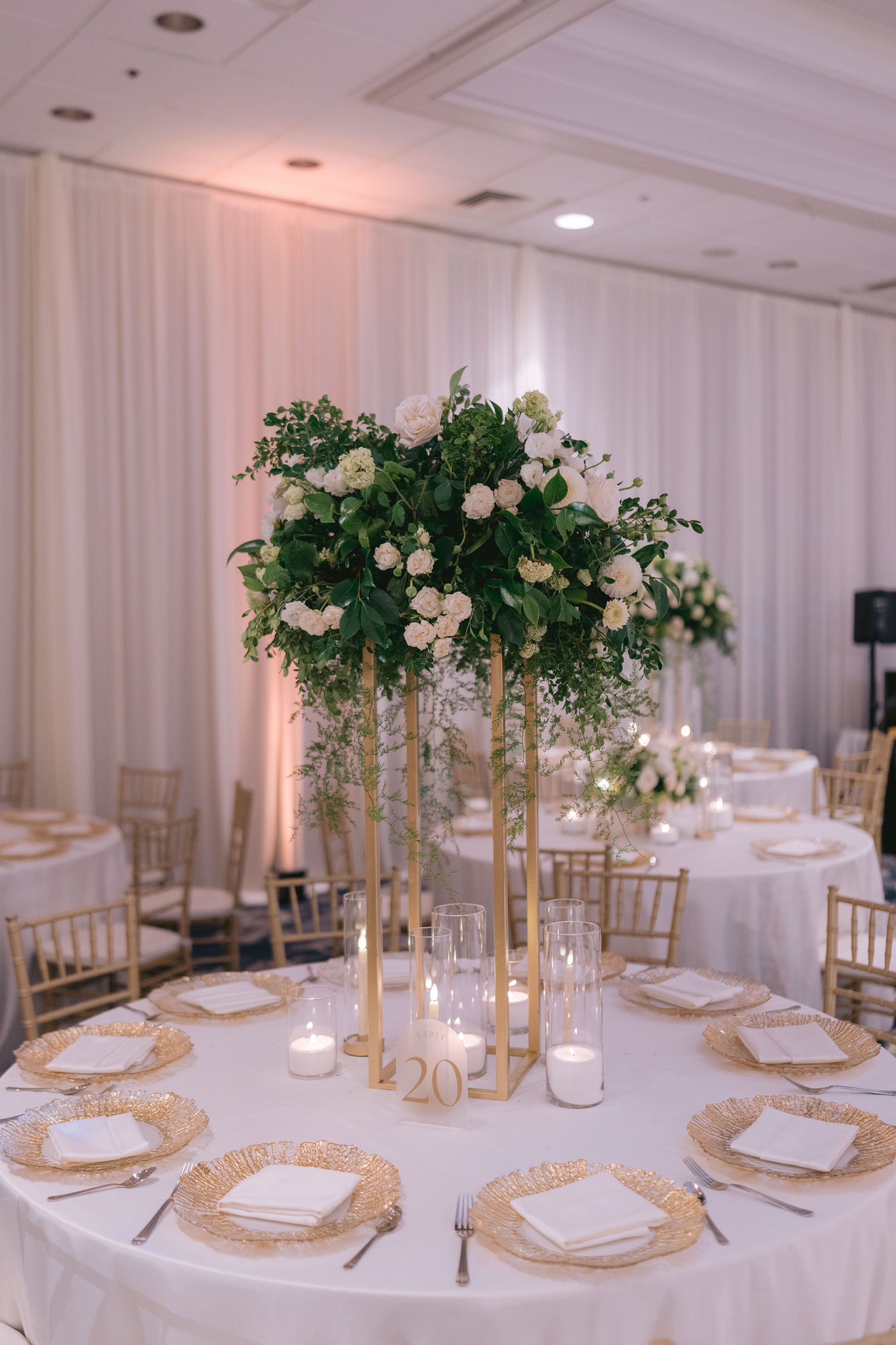 A round banquet table set for a formal event with white tablecloths, gold charger plates, white napkins, and silverware. The centerpiece is a tall floral arrangement of white flowers and green foliage on a gold stand. There are candles and a table nu