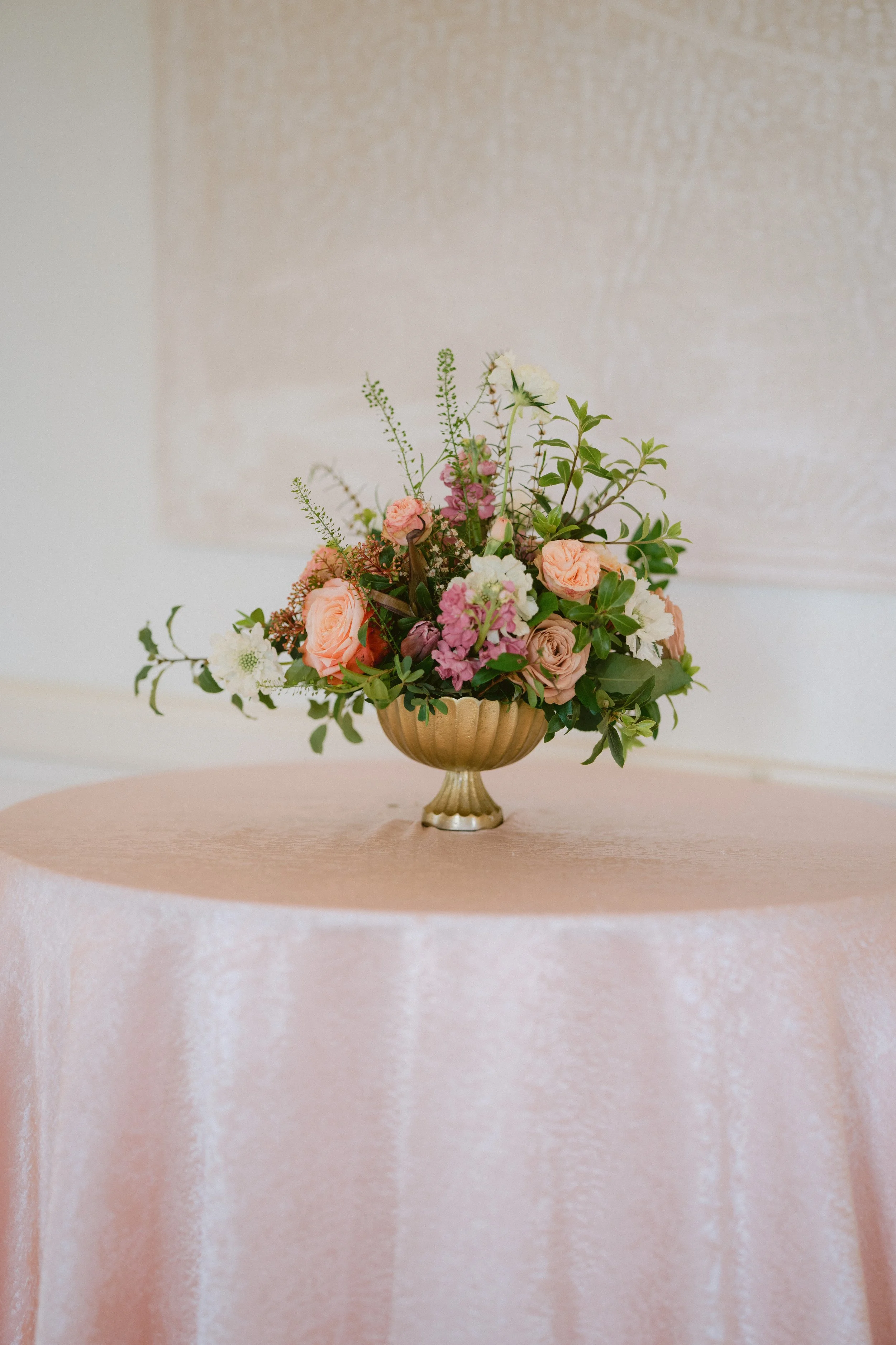 A floral arrangement with pink, white, and peach flowers, and green leaves in a gold vase on a pink tablecloth.