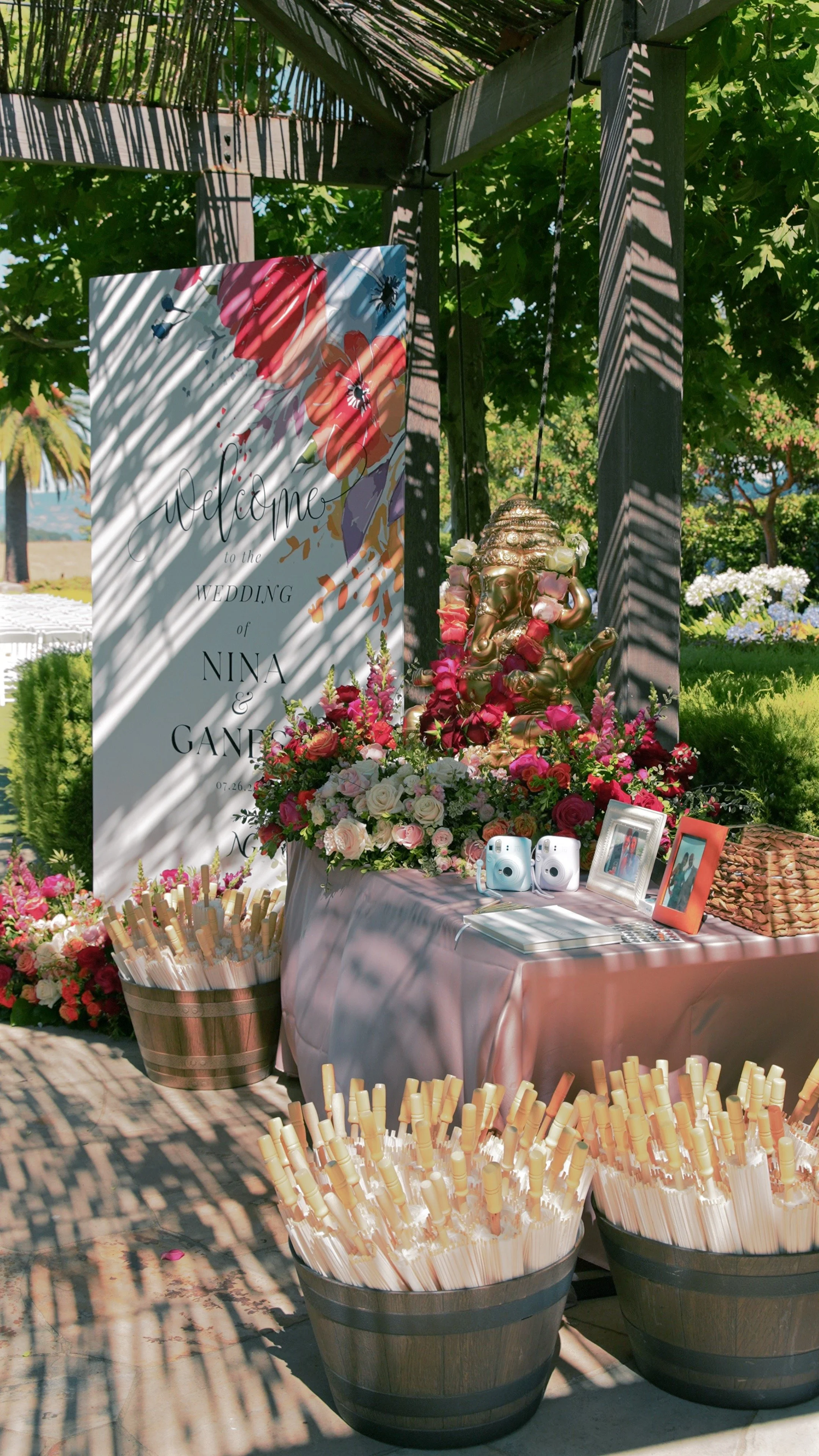 Outdoor wedding reception area decorated with pink and white flowers, a large welcome sign, a golden Ganesha statue surrounded by flowers, framed photos, and two Polaroid cameras on a table. Baskets of white rolled-up napkins or paper straws are in t