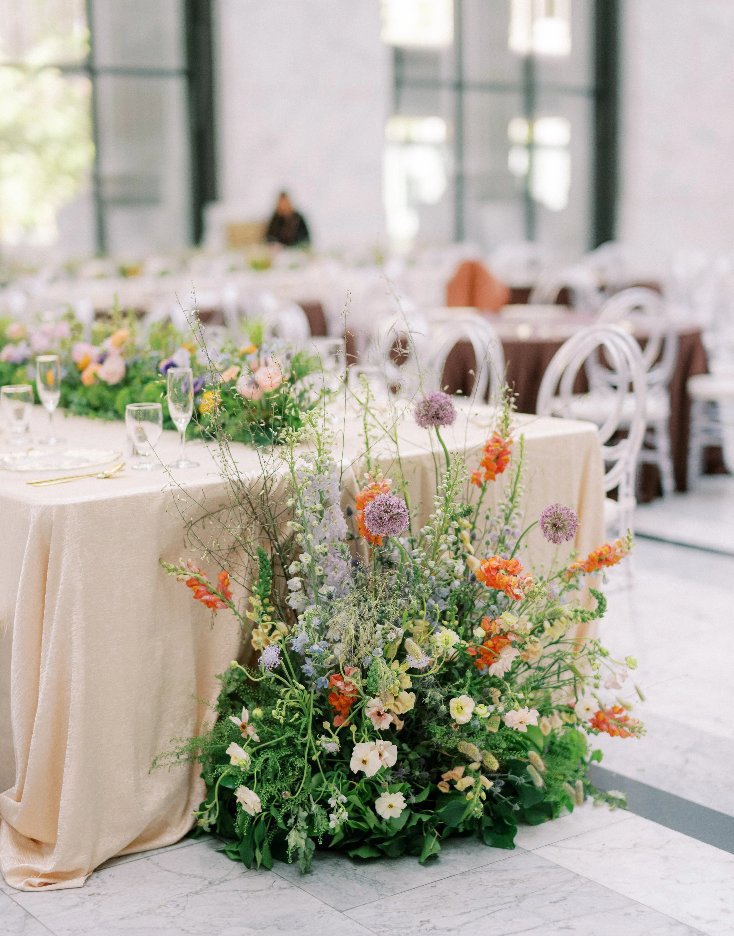 Table decorated with colorful flowers in a vase, set for a formal event in a bright room with large windows and white chairs.