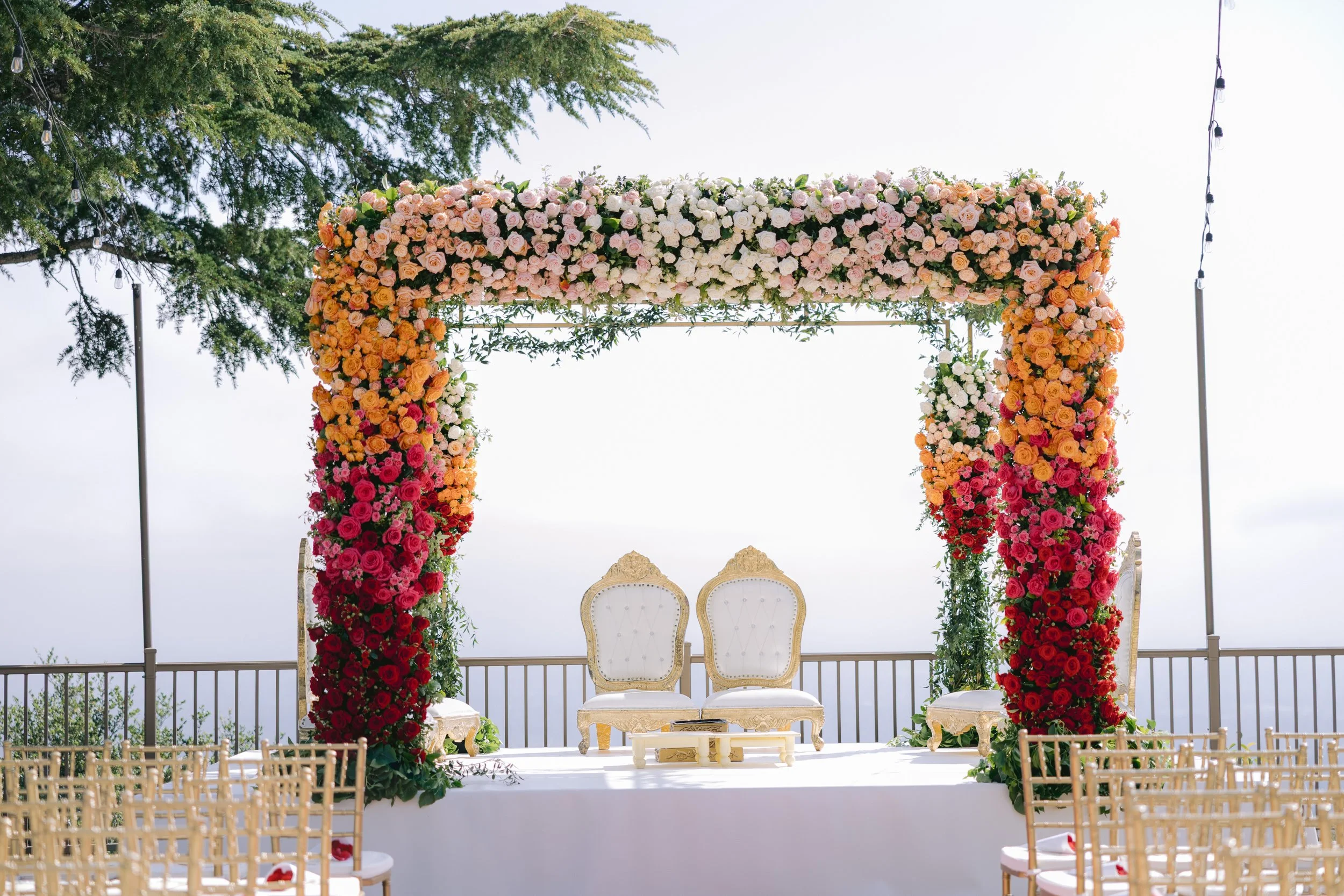 A wedding arch decorated with an abundance of pink, white, peach, orange, and red roses, with two ornate white chairs underneath. The setting is outdoors with a tree and string lights in the background, overlooking a railing and an overcast sky.