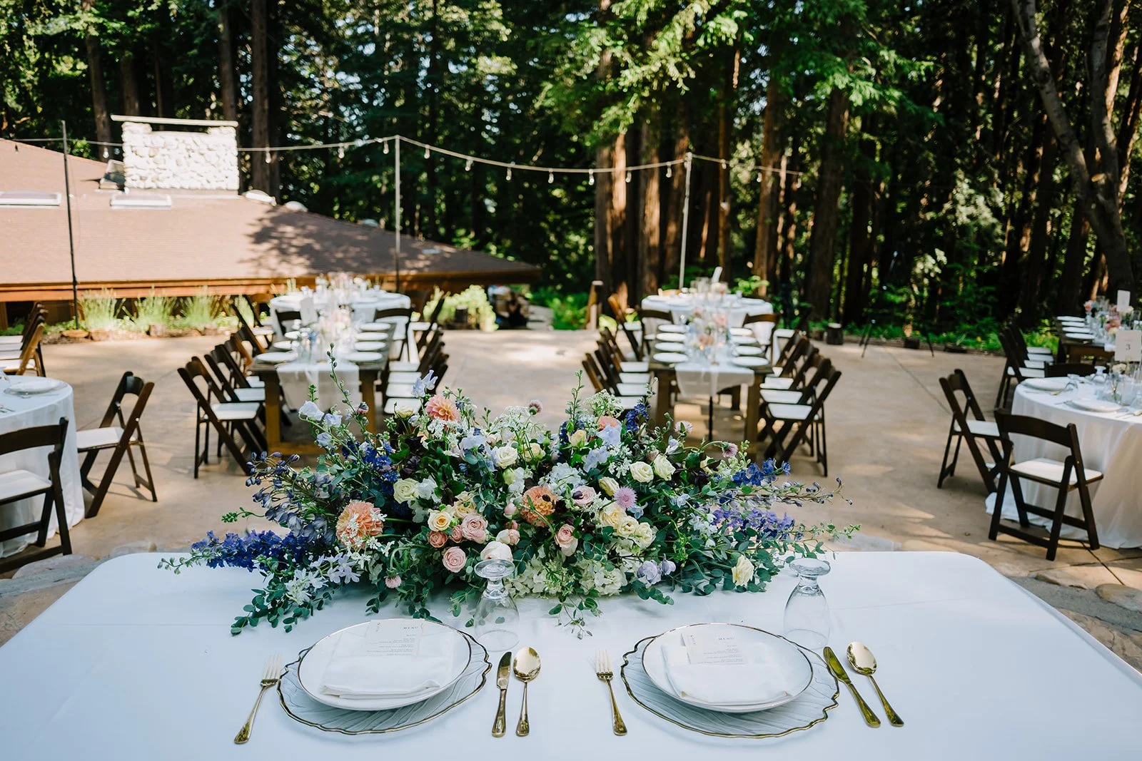 Wedding reception setup outdoors with round tables, white tablecloths, elegant tableware, and a large colorful floral centerpiece, surrounded by black chairs, with trees and string lights in the background.