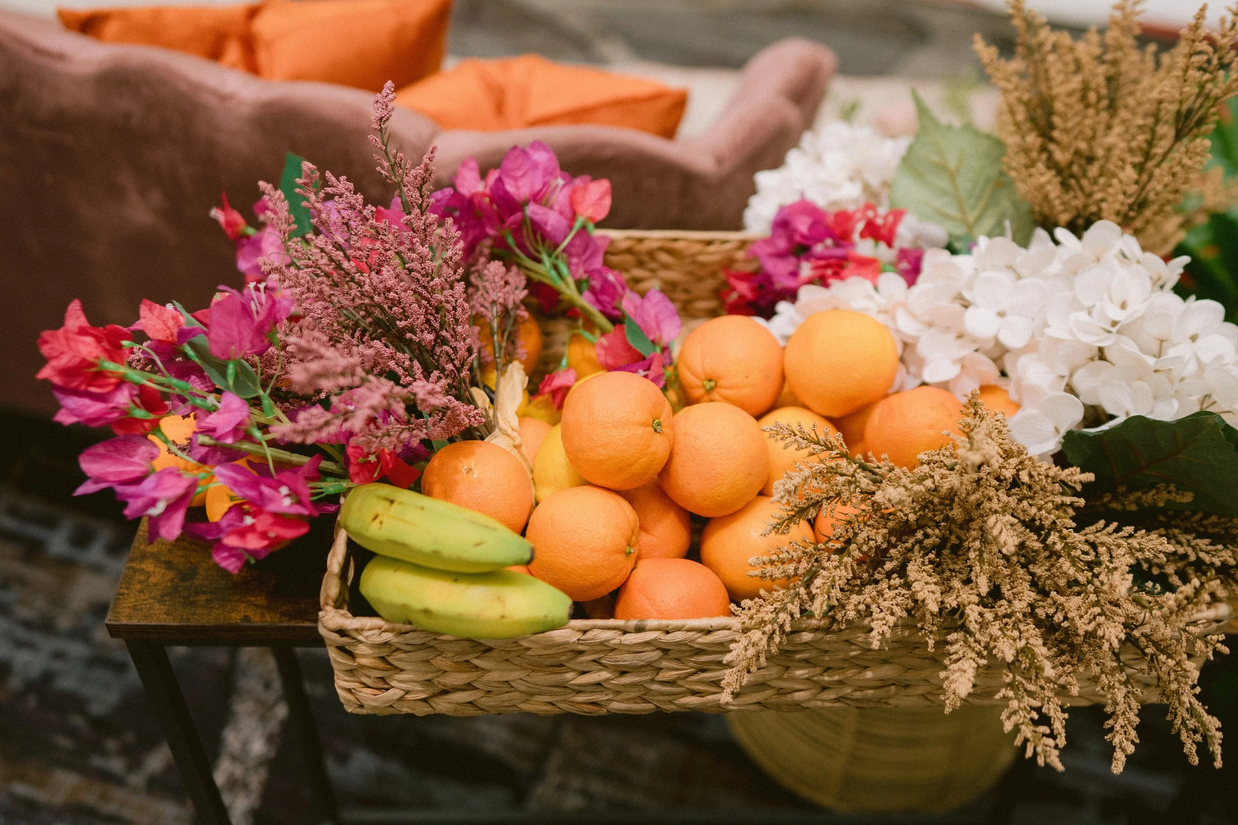 A wicker basket filled with oranges, bananas, and various colorful flowers, sitting on a wooden surface.