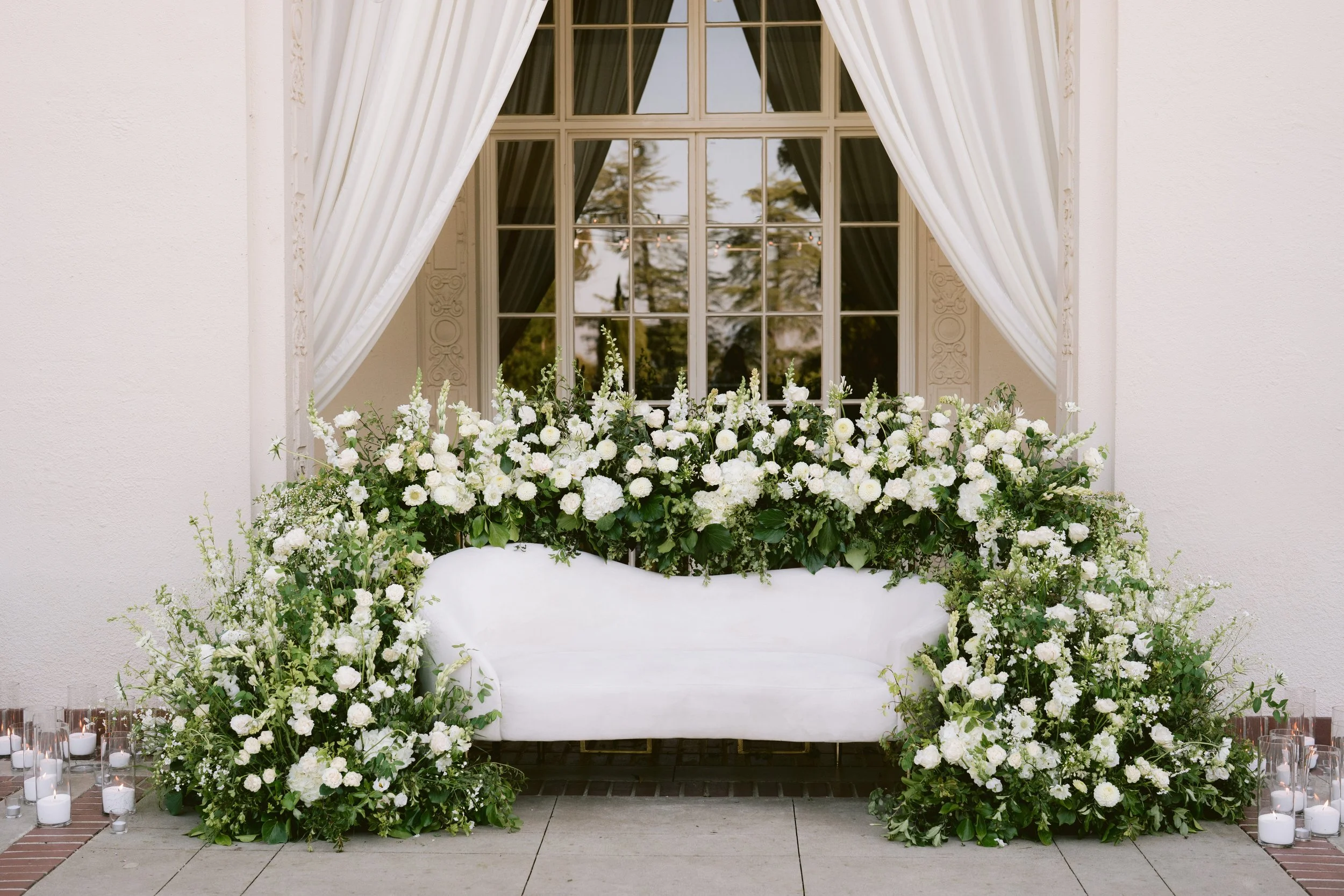 A floral arrangement on a white sofa in front of a window with white curtains. Candles are on the ground around the sofa.