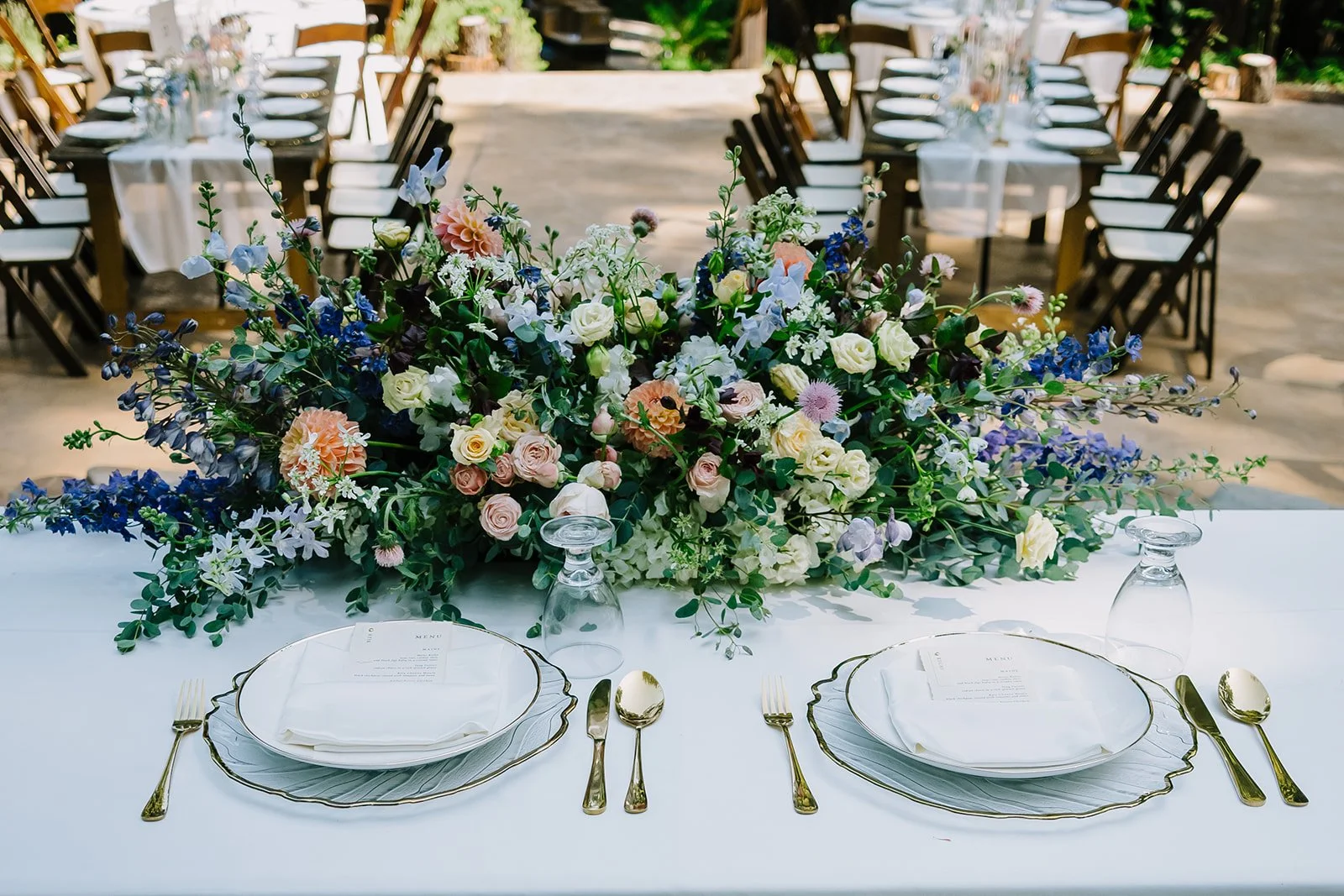 Table set for an outdoor wedding with a large floral centerpiece, white plates, gold utensils, and upside-down glasses on a white tablecloth.