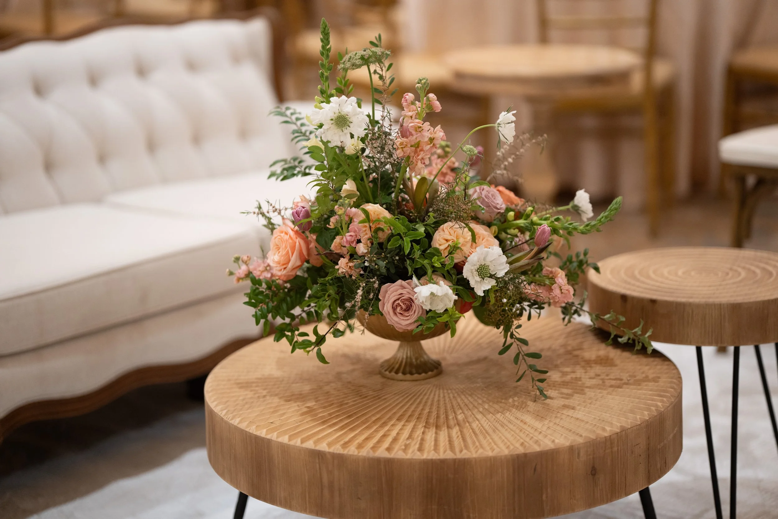 A floral arrangement in a wooden pedestal vase on a round wooden table, with a white tufted sofa and wooden chairs in the background.