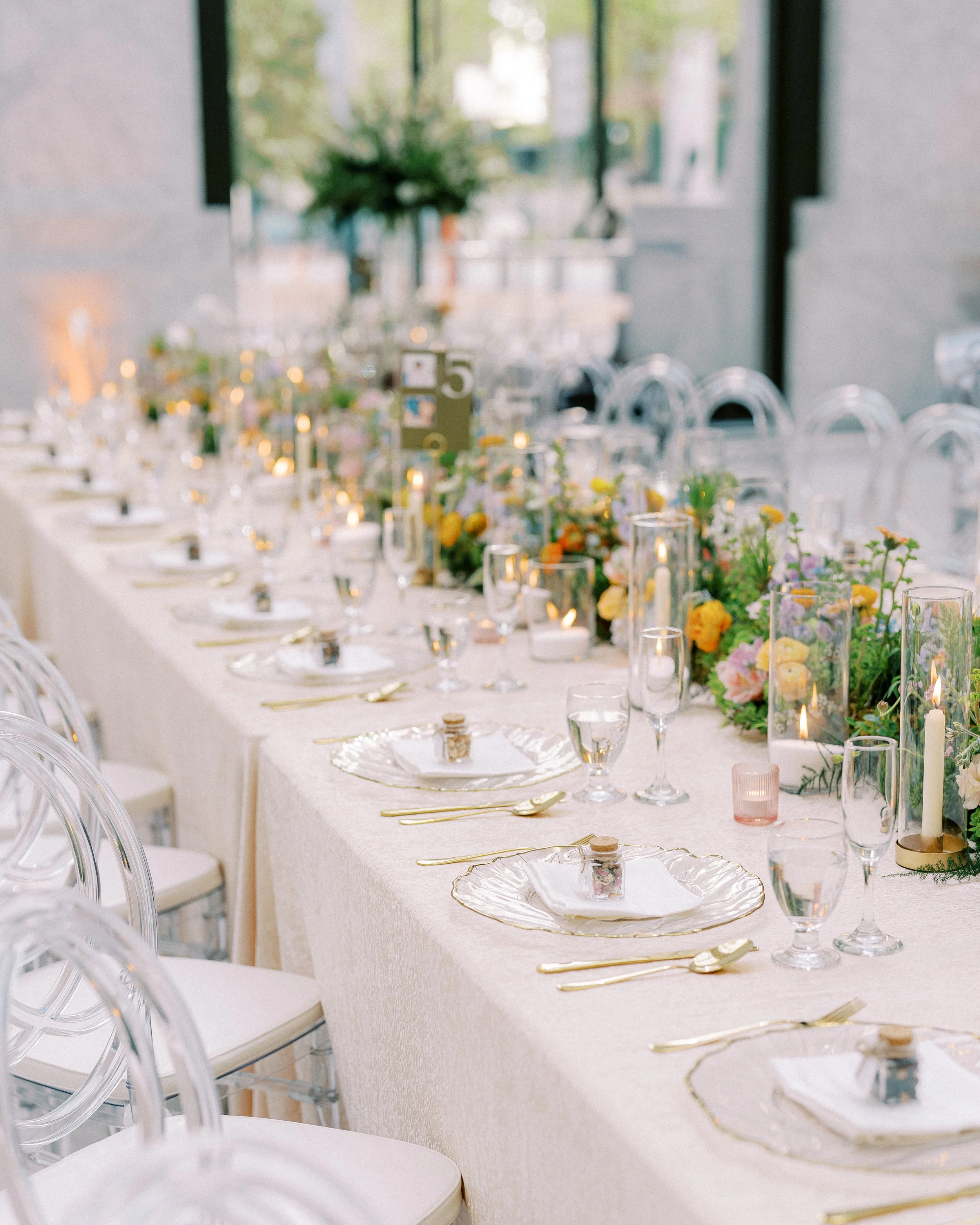 Elegant banquet table set with glassware, gold cutlery, decorative plates, floral arrangements, candles, and a white tablecloth, with a large window in the background letting in natural light.