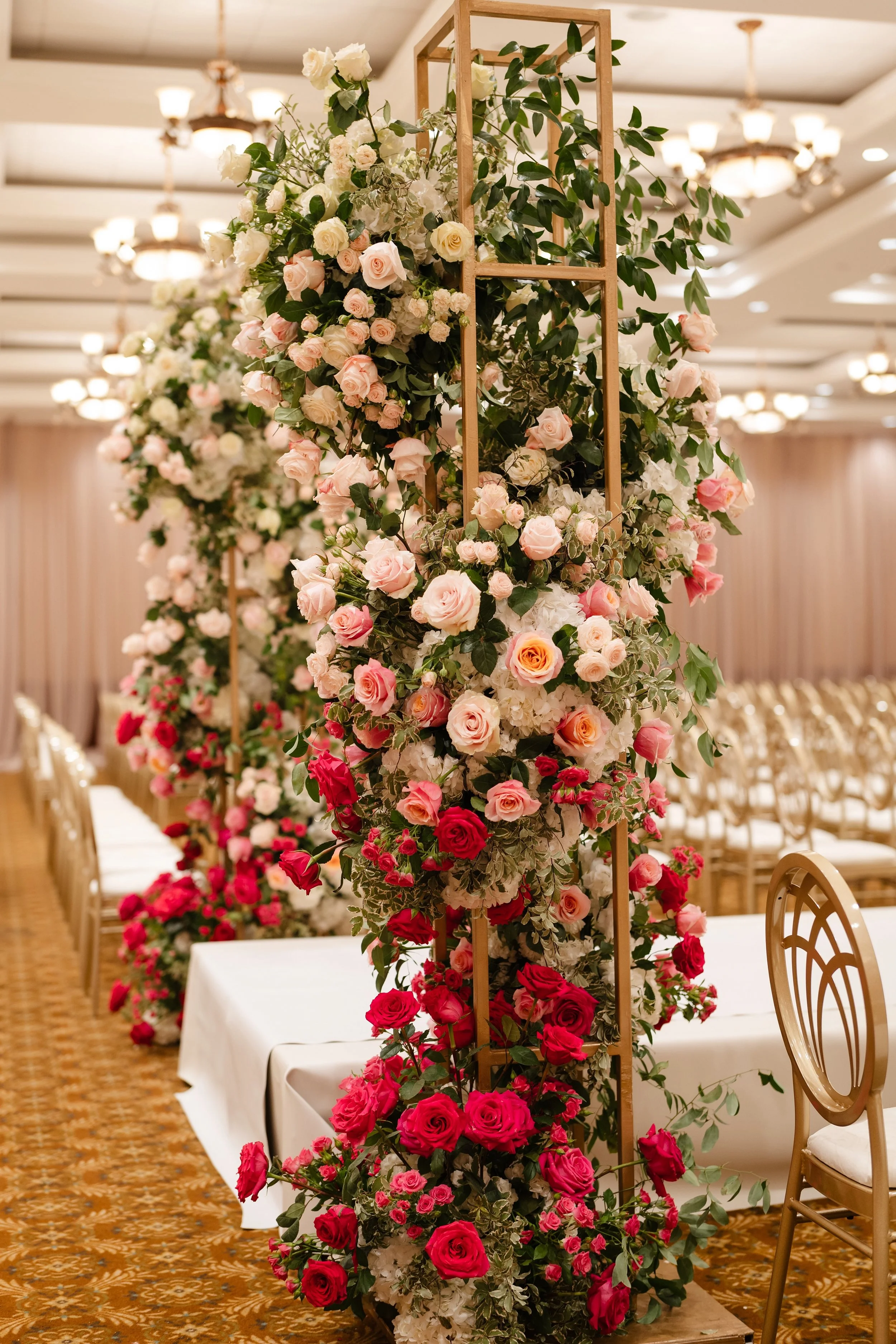A floral decoration with pink and white roses on a wooden structure in a banquet hall decorated with chandeliers and curtains.