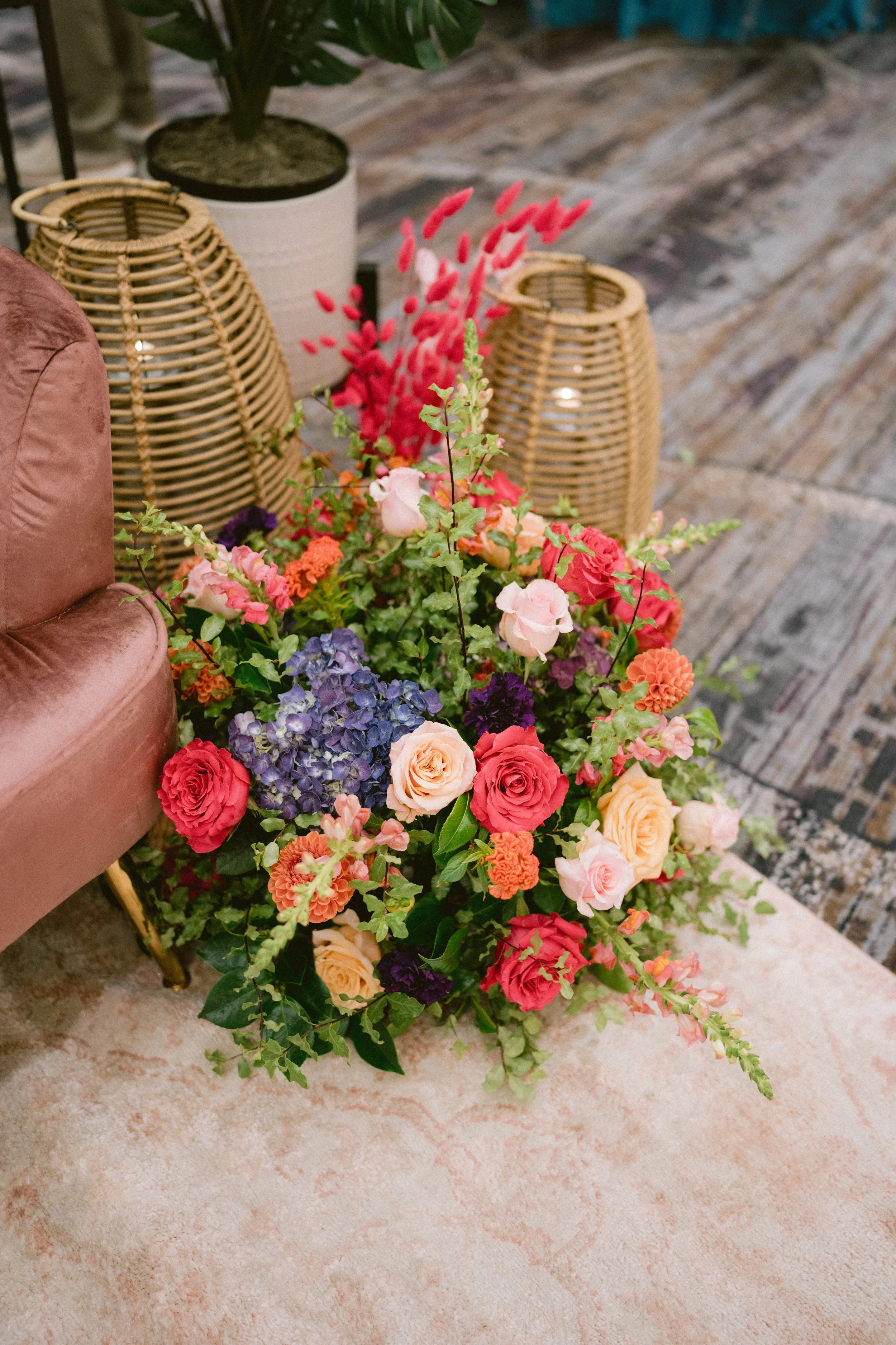 Colorful bouquet of various flowers including roses, hydrangeas, and others, placed on a marble floor near rattan lanterns and a pink velvet chair, with some indoor greenery and textured flooring in the background.