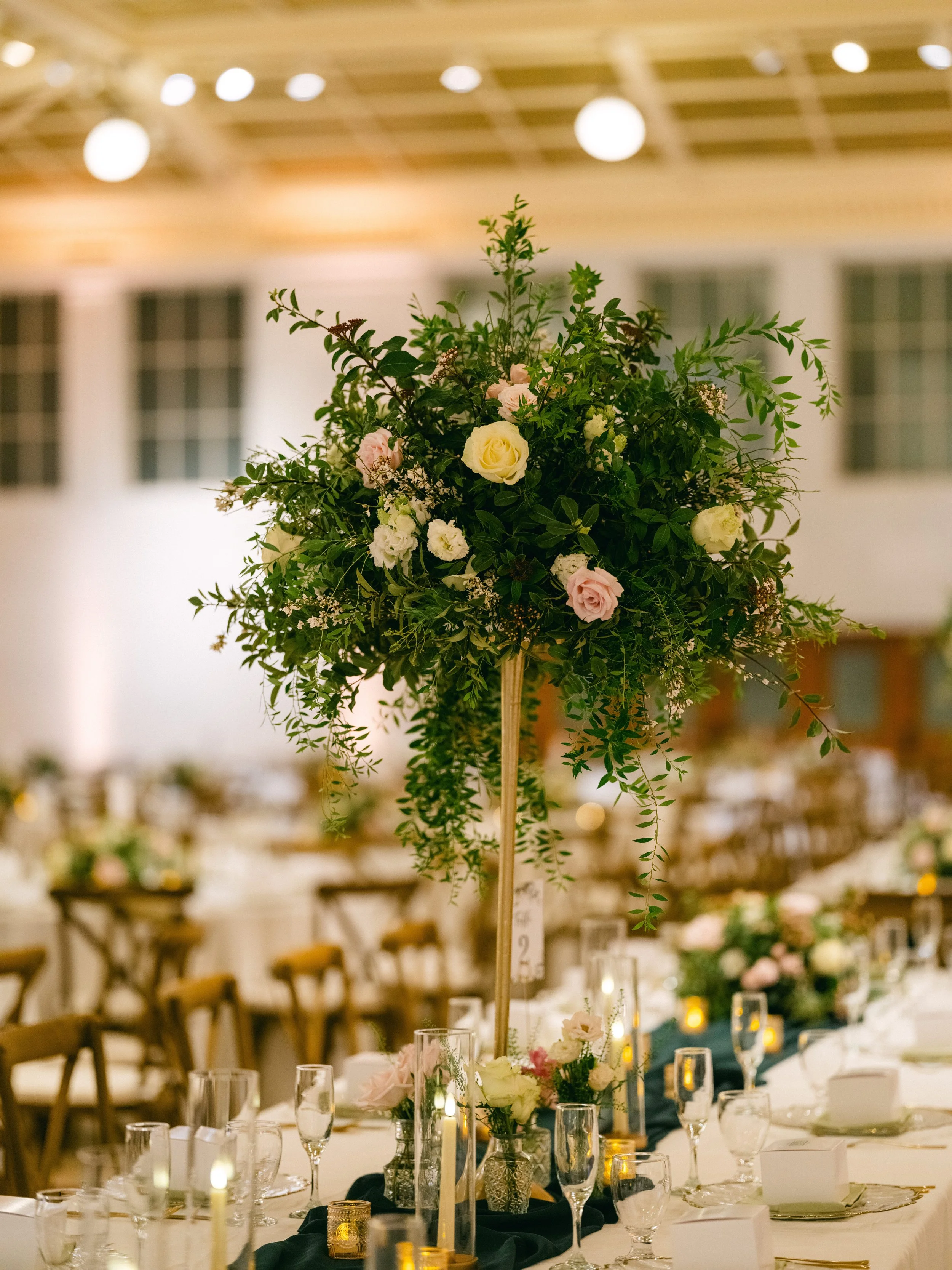 A tall floral centerpiece with pink and white roses and assorted greenery on a decorated banquet table in a banquet hall.