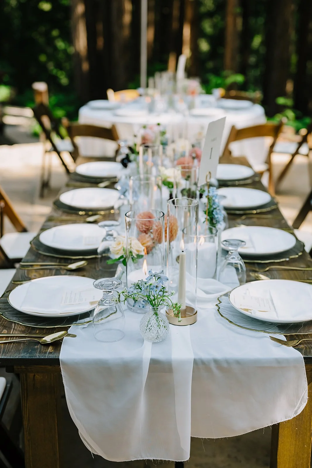 Outdoor wedding reception table decorated with white tablecloth, glass vases with flowers, candles, and place settings with white plates, gold utensils, and upside-down glasses, surrounded by wooden chairs.