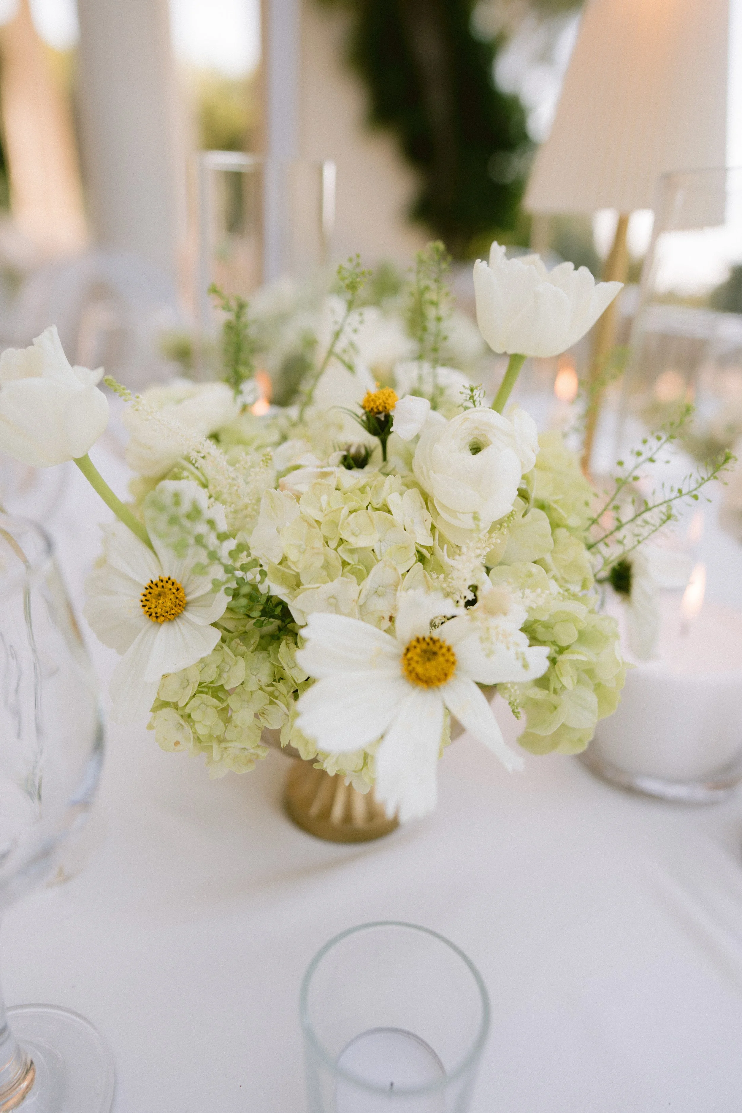 A bouquet of white and cream-colored flowers, including daisies and other blooms, in a small vase on a table with a white tablecloth, surrounded by empty clear glass glasses.