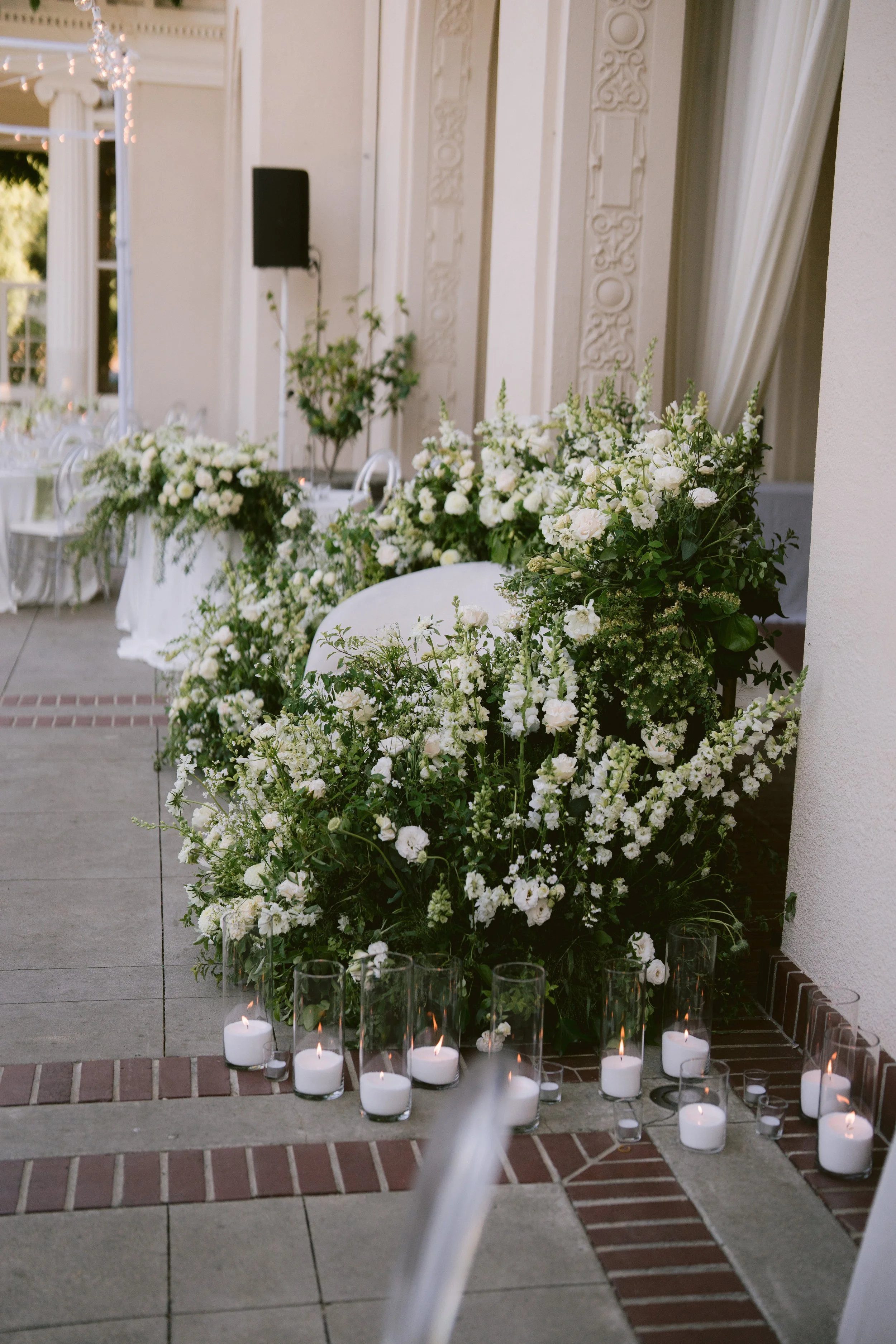 A decorative floral arrangement of white flowers and greenery on a table, with lit candles in glass holders arranged on the floor nearby, at an indoor or outdoor event venue with ornate white walls and columns.