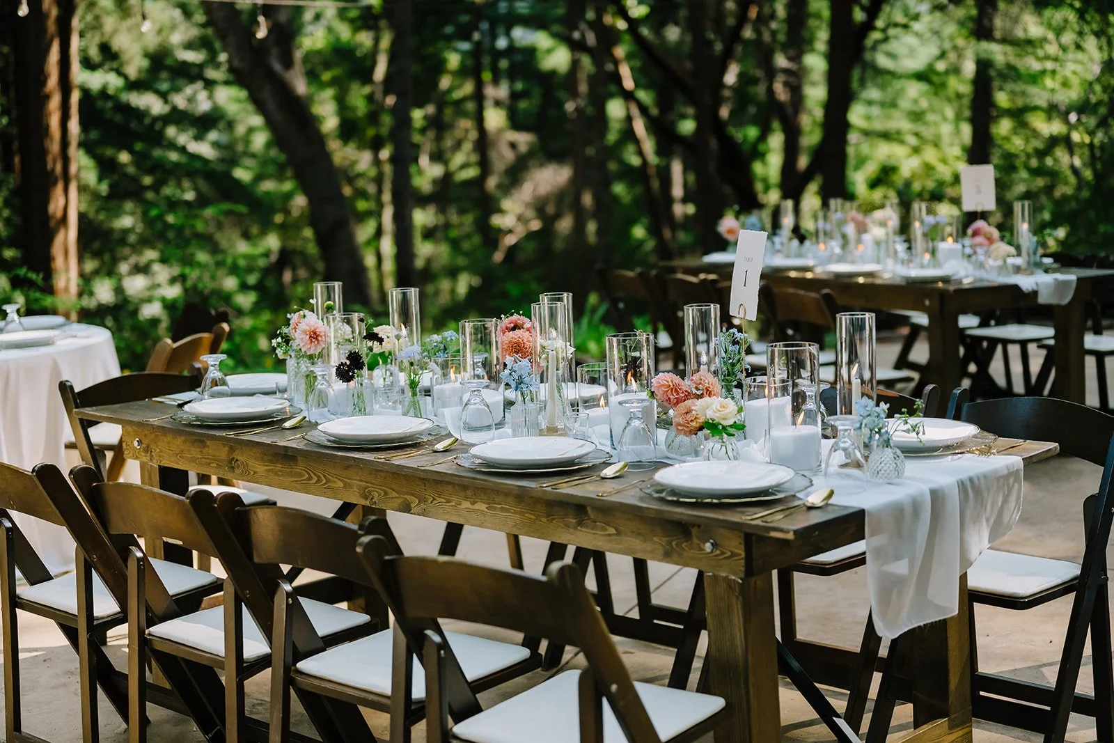 Outdoor dining setup with long wooden table, white tablecloth, plates, gold utensils, glassware, and floral centerpieces, surrounded by chairs, in a wooded area.