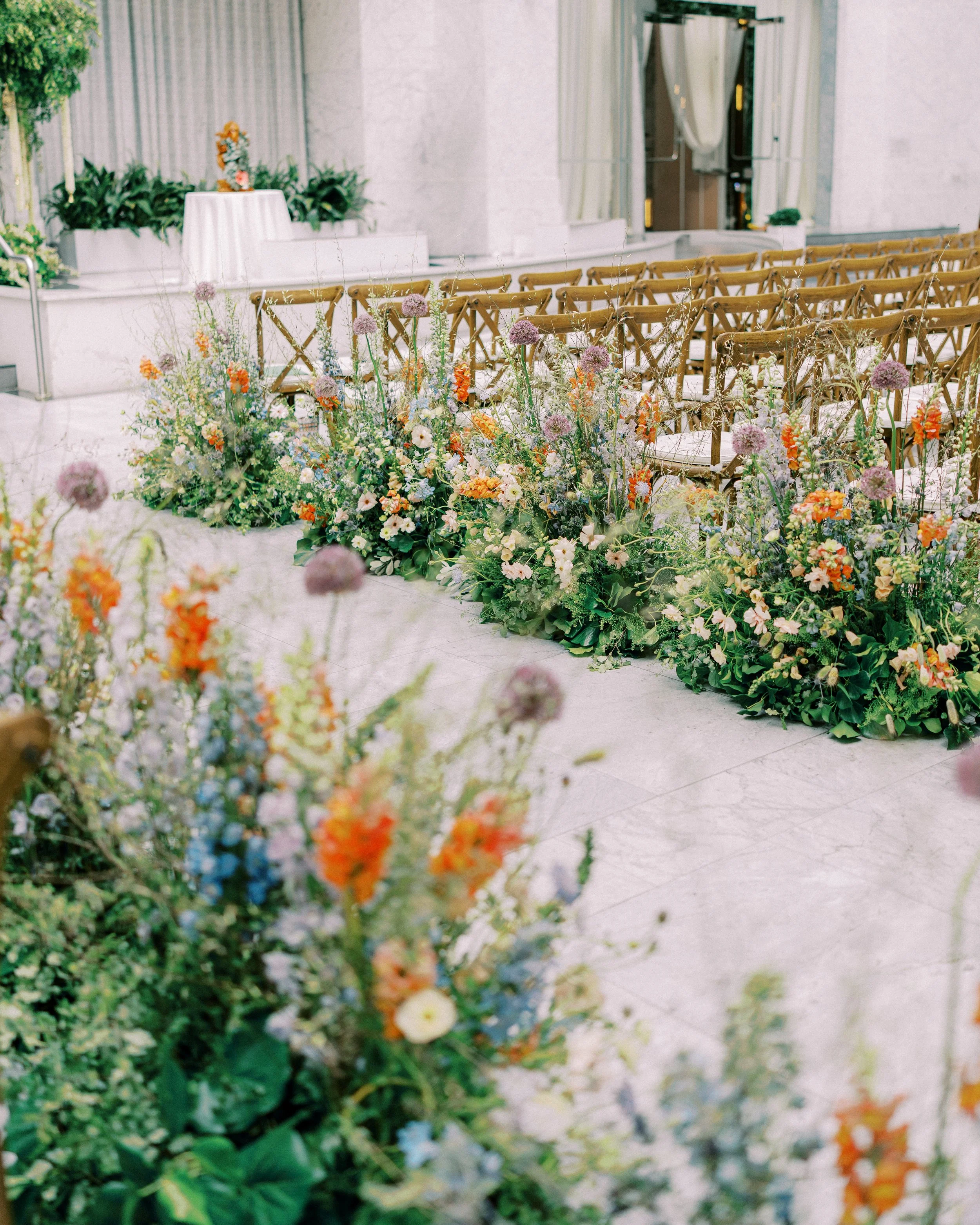 Indoor setting with wooden chairs arranged in rows, adorned with a lush display of various flowers in front. A small, elevated stage with a white tablecloth and a floral arrangement is at the front, with greenery and potted plants in the background.