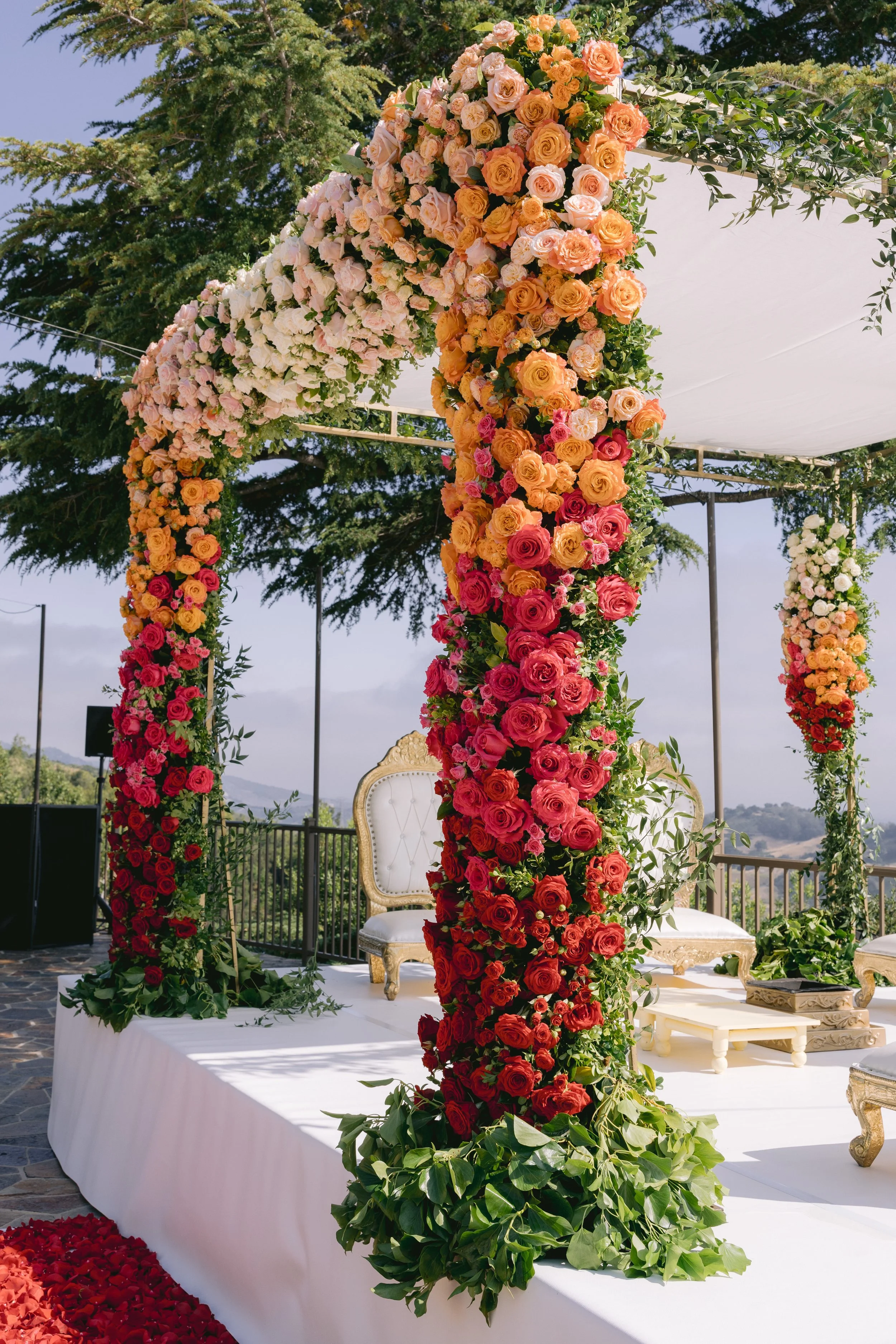 A floral wedding arch decorated with a variety of orange, pink, white, and red roses, with greenery, set outdoors on a white platform under a white canopy, with trees and a wooden railing in the background.