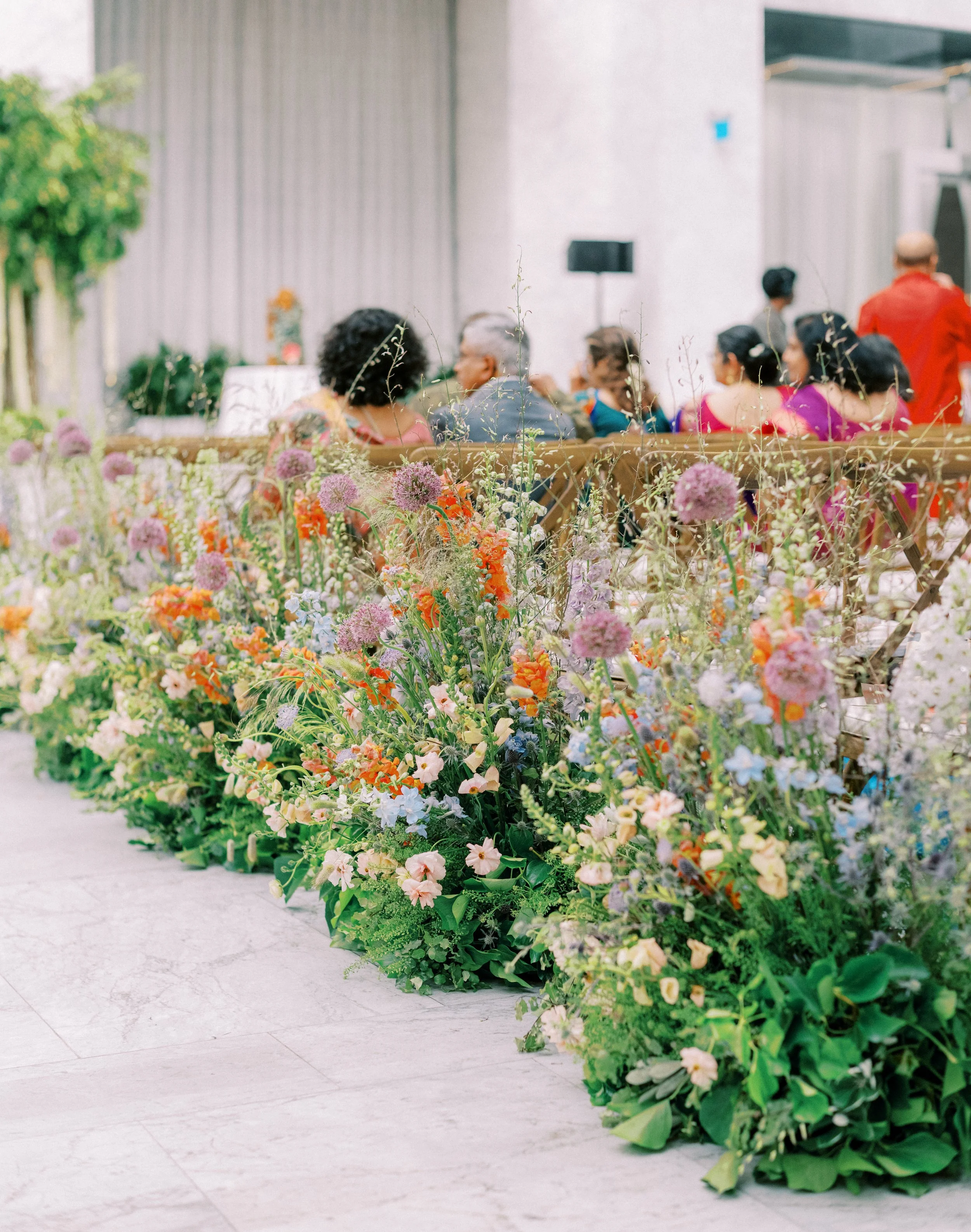 Colorful flower arrangements placed at the edge of an indoor space with people seated in the background.