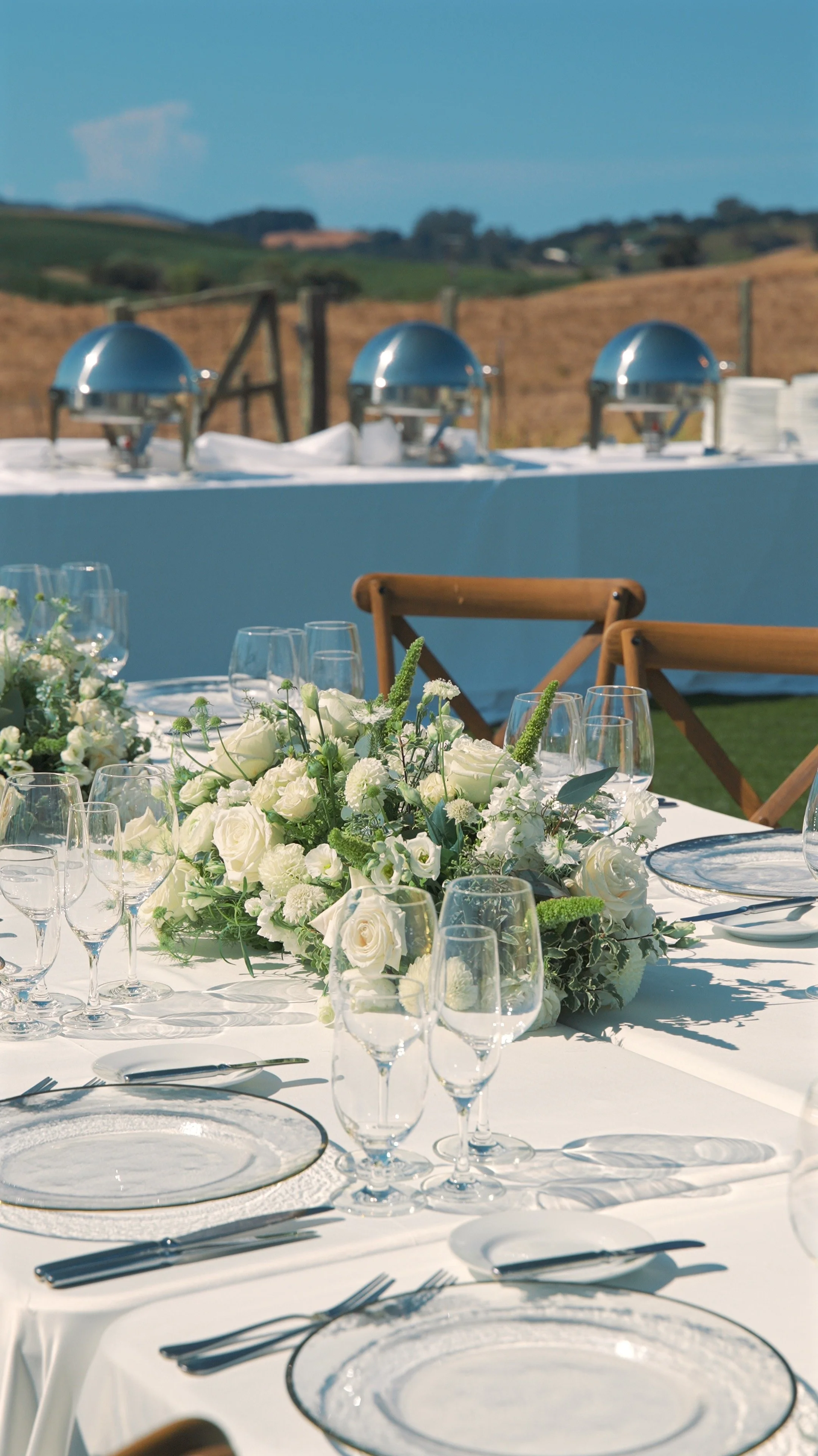 Elegant outdoor dining table setup with white tablecloth, floral centerpiece, and neatly arranged glassware and silverware.