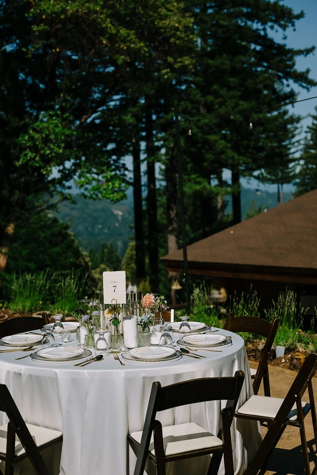 Round outdoor table set for a formal event with white tablecloth, plates, silverware, glasses, and centerpieces of flowers and candles, surrounded by black chairs with white cushions, with trees and a building with a sloped roof in the background.