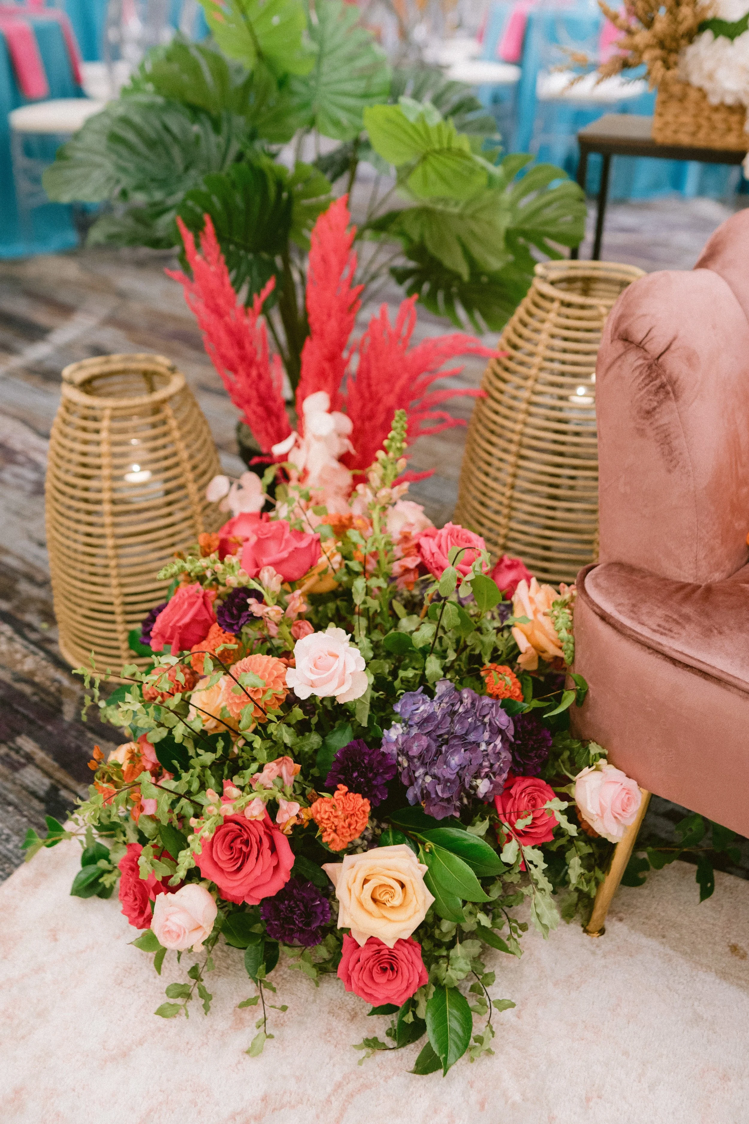 Colorful floral arrangement with pink, purple, orange, and cream roses, along with greenery, placed on a light-colored carpet. In the background, there are large leafy plants and decorative woven lampshades, and a pink velvet chair is partially visib