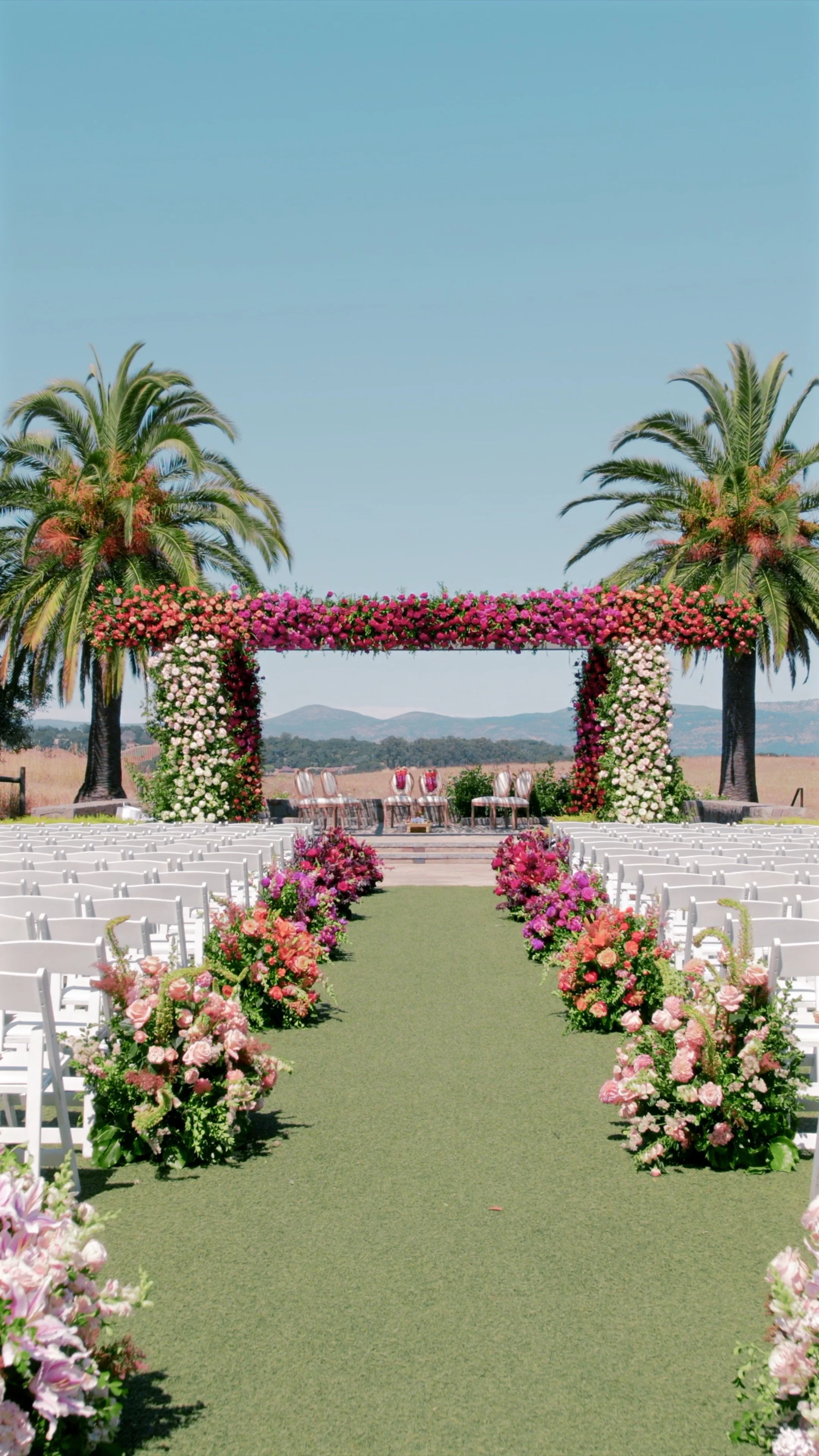 A beautifully decorated outdoor wedding venue with a floral arch, palm trees, and rows of white chairs on both sides of a green aisle leading to a stage with chairs and a table, set against a rural landscape with hills and blue sky.