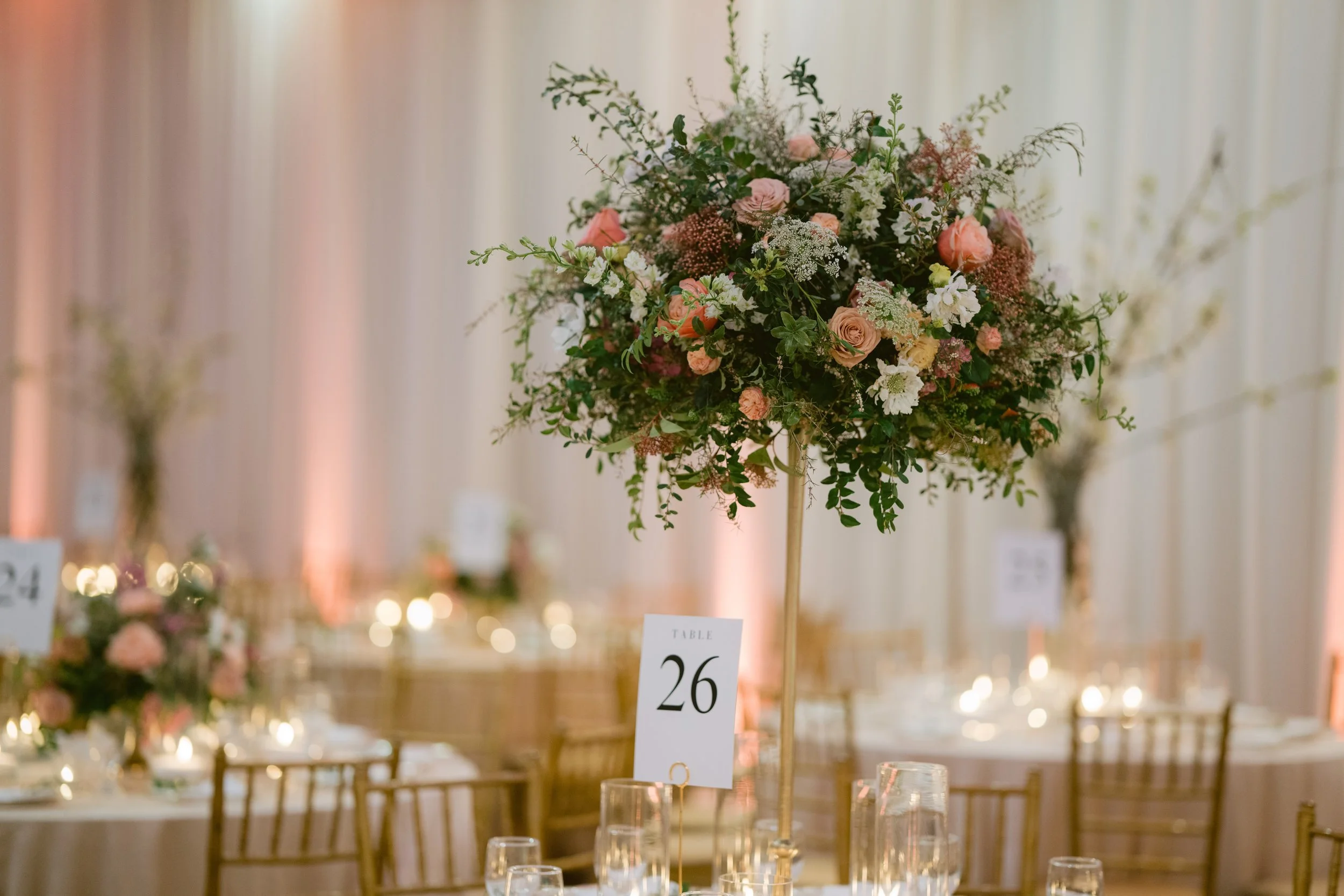 A tall floral centerpiece with pink and white roses, white and pink flowers, and green foliage on a table at an event.
