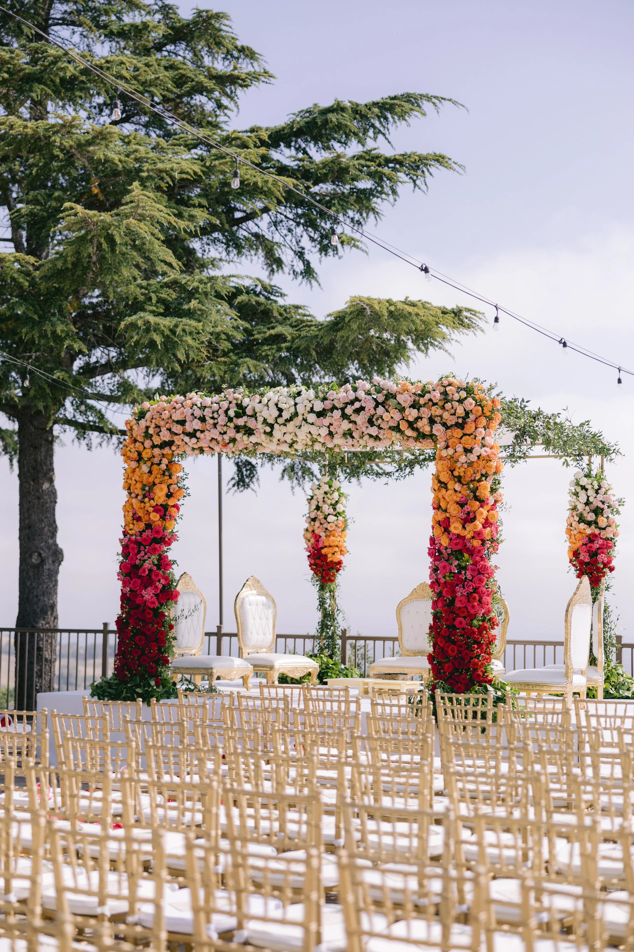 Outdoor wedding ceremony setup with white chairs, a floral arch with pink, orange, and white roses, and string lights hanging from trees against a clear sky.