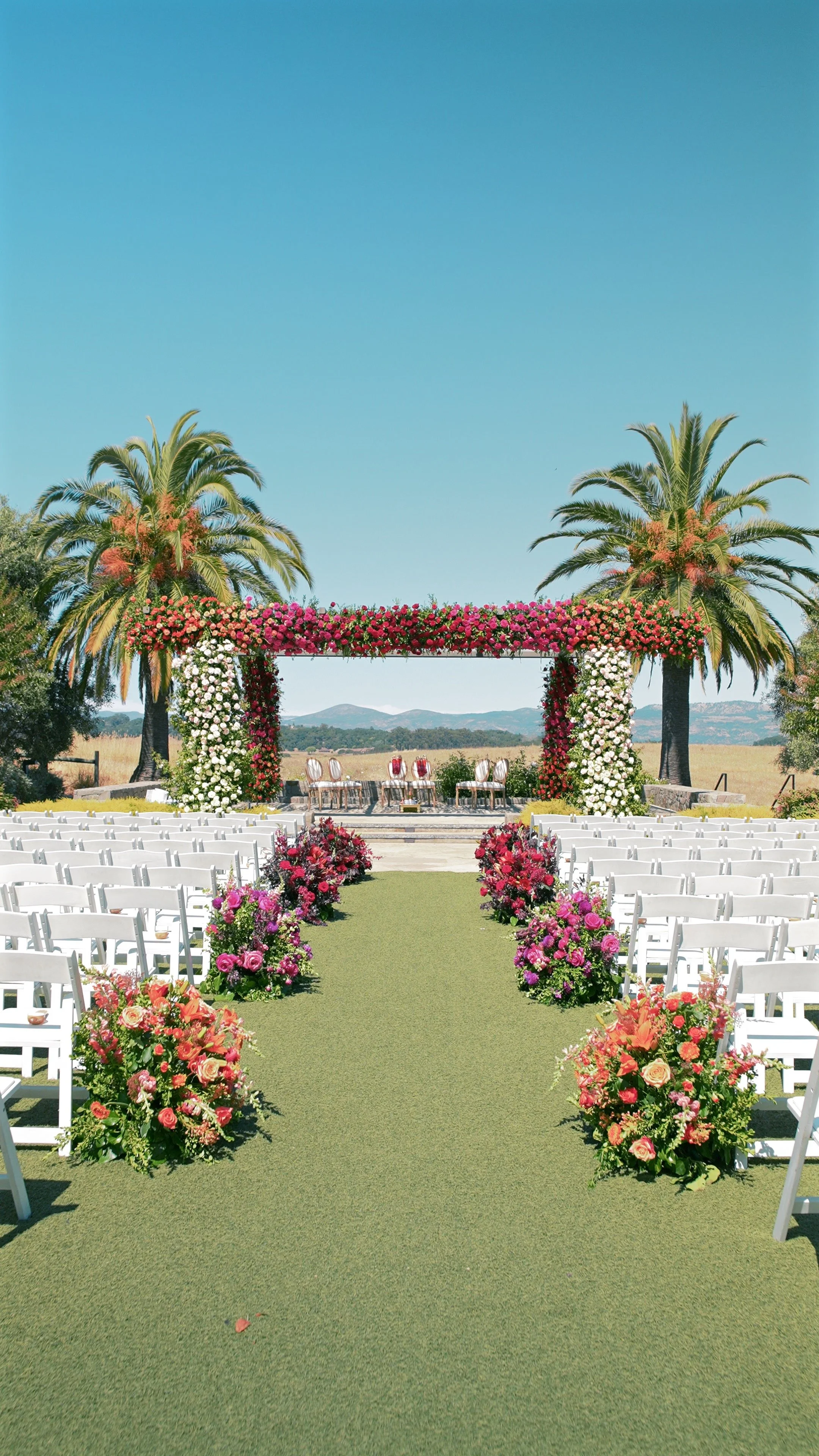 Outdoor wedding setup with white chairs, floral arrangements, and an arch decorated with pink and white flowers under palm trees on a sunny day.