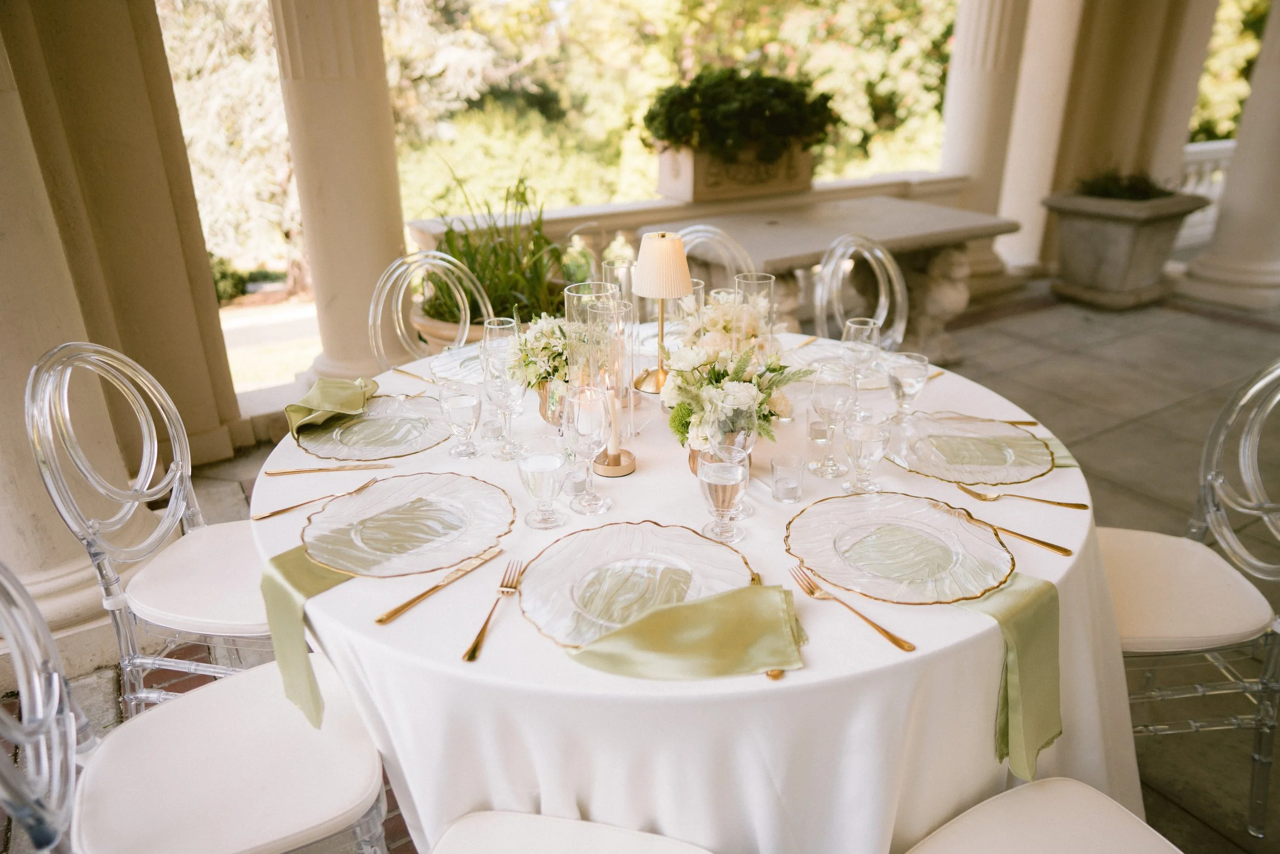 Formal dining table set with glassware, white floral centerpiece, and a small lamp, with a background of windows and greenery.