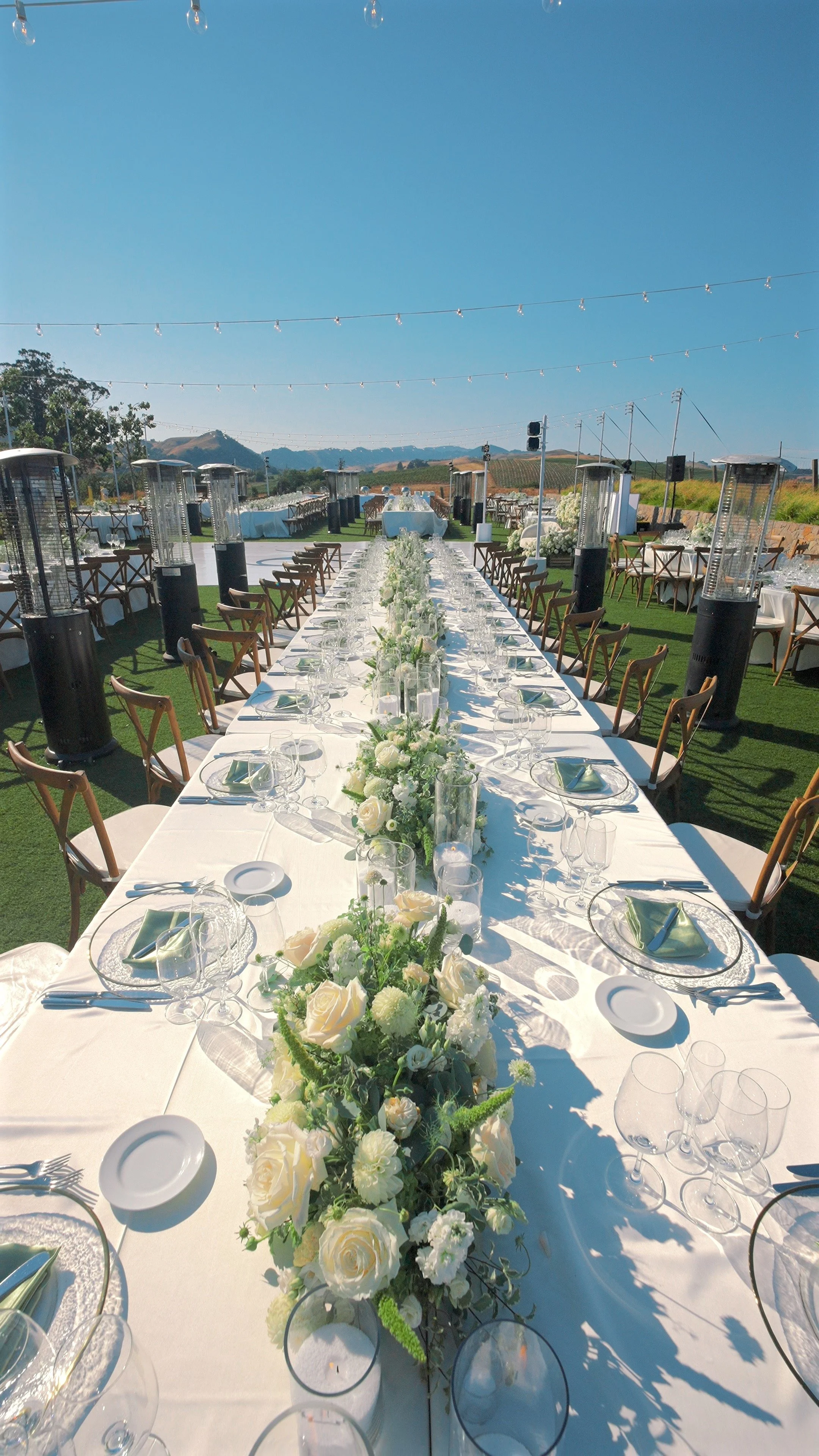 Outdoor banquet setup with long table decorated with white flowers and tableware, under string lights, with mountains in the background.