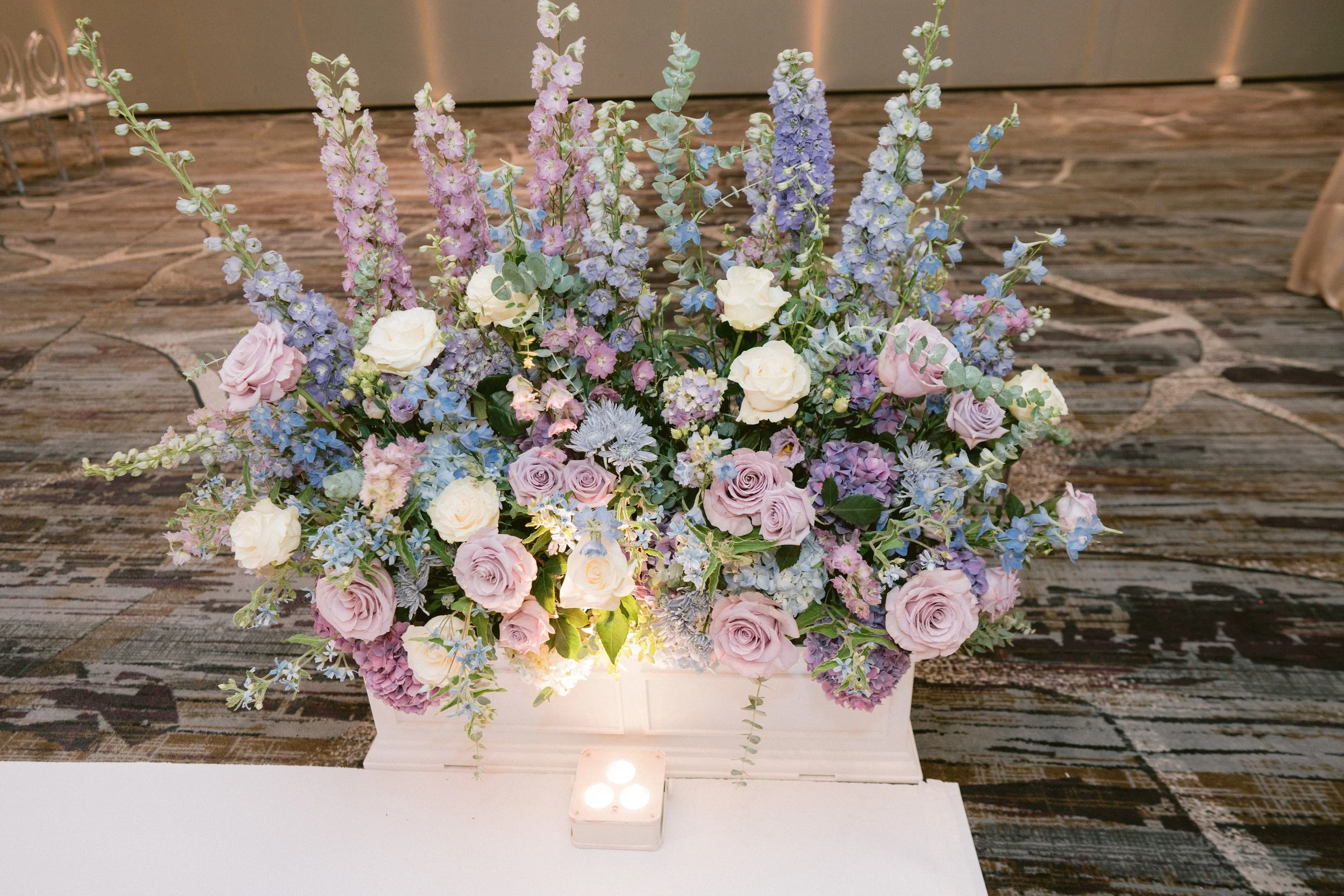 A large bouquet of pastel-colored flowers, including roses, delphiniums, and other blossoms, arranged in a white rectangular container on a table.